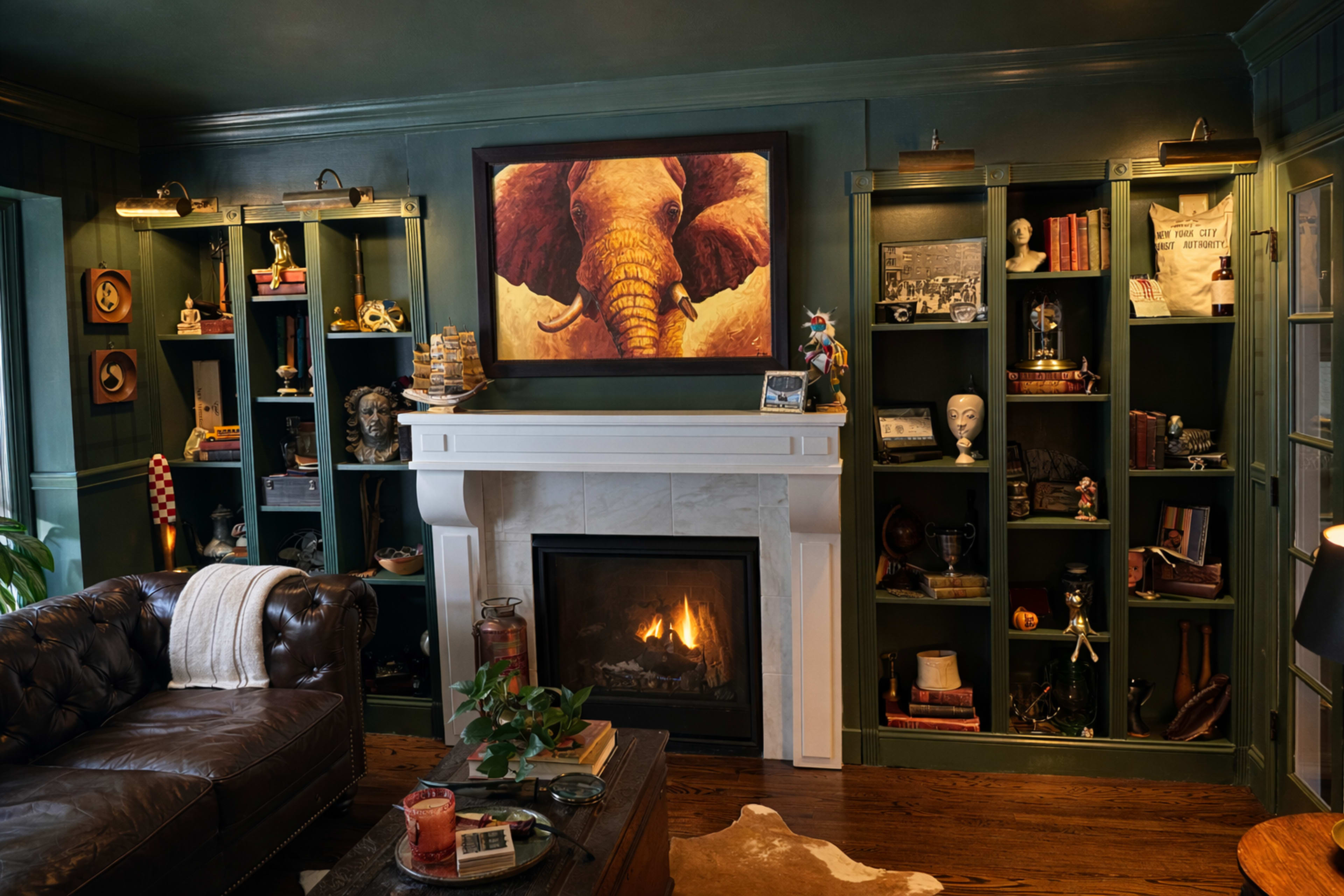 The image shows a cozy living room with a leather couch, a fireplace with a marble surround, and shelves filled with various decorative items and books.
