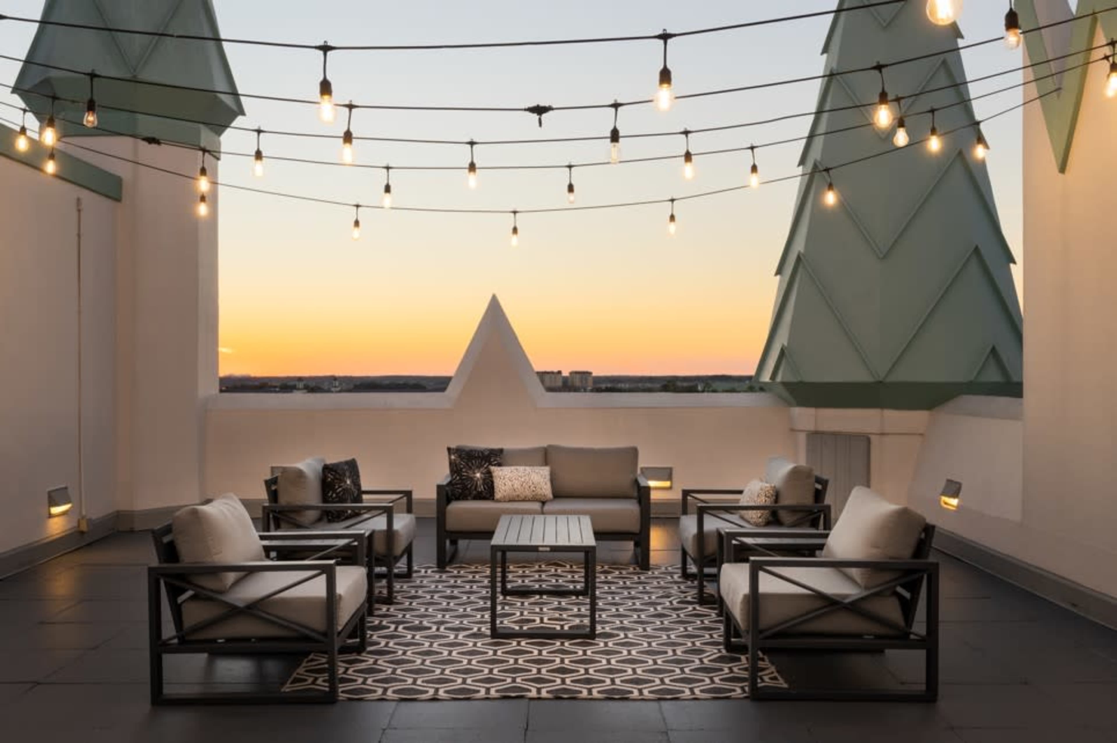 A rooftop terrace features a seating area with a sofa, armchairs, and a coffee table, surrounded by string lights and a colorful sunset in the background.