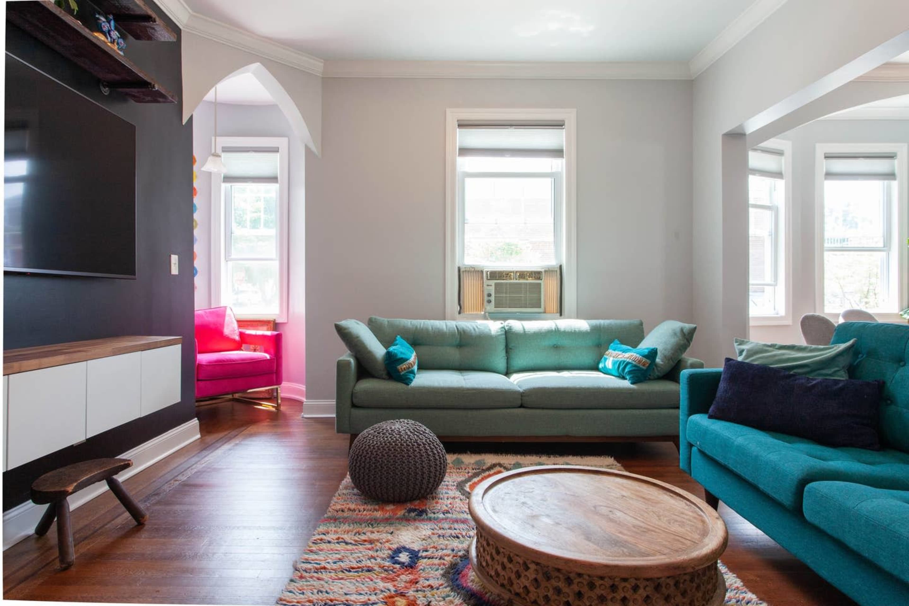 The image shows a living room featuring two sofas, a coffee table, and a window air conditioning unit, with natural light coming through the windows.