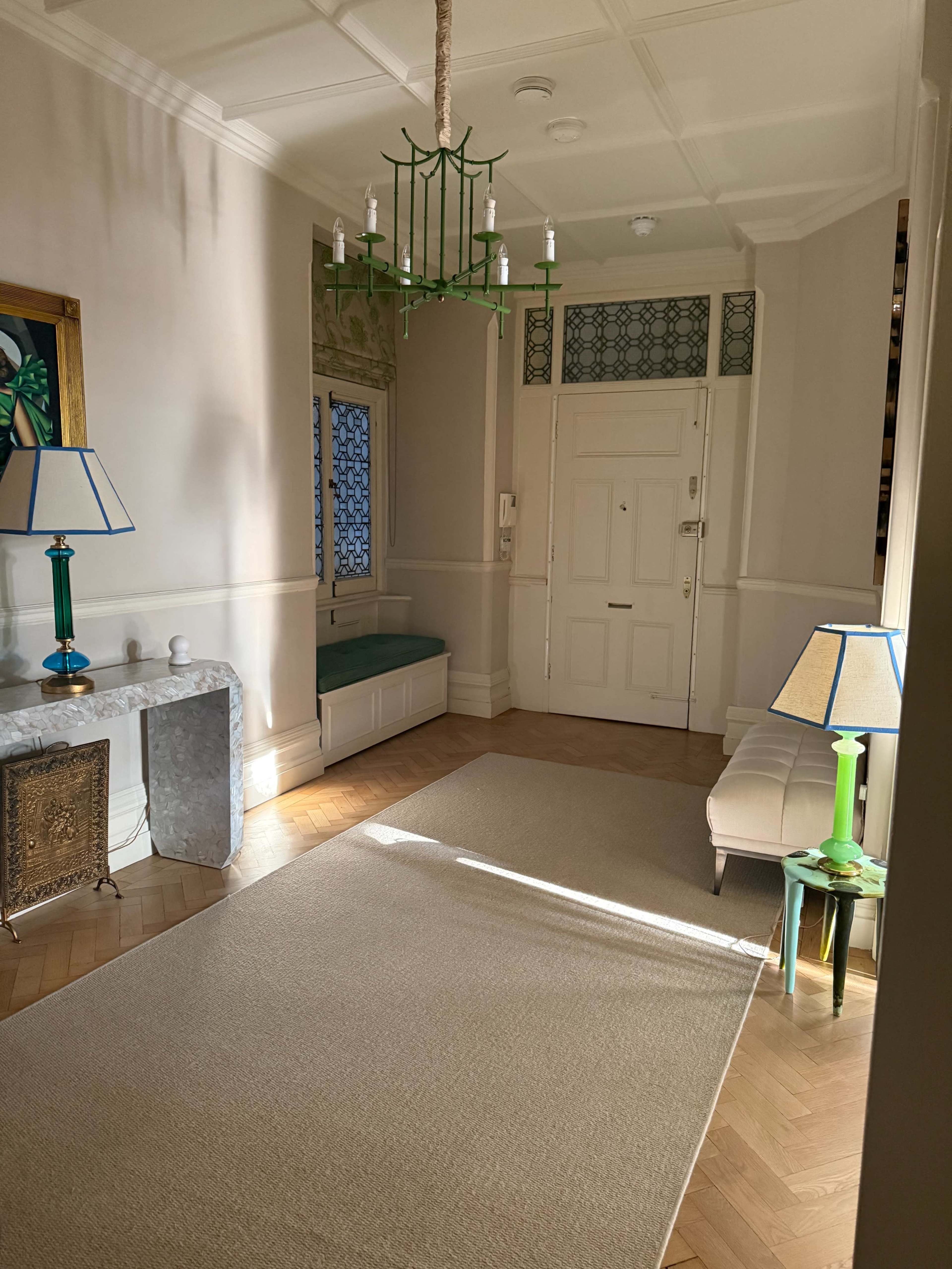 The image shows a well-lit hallway with a decorative chandelier, green accents, and a small seating area.