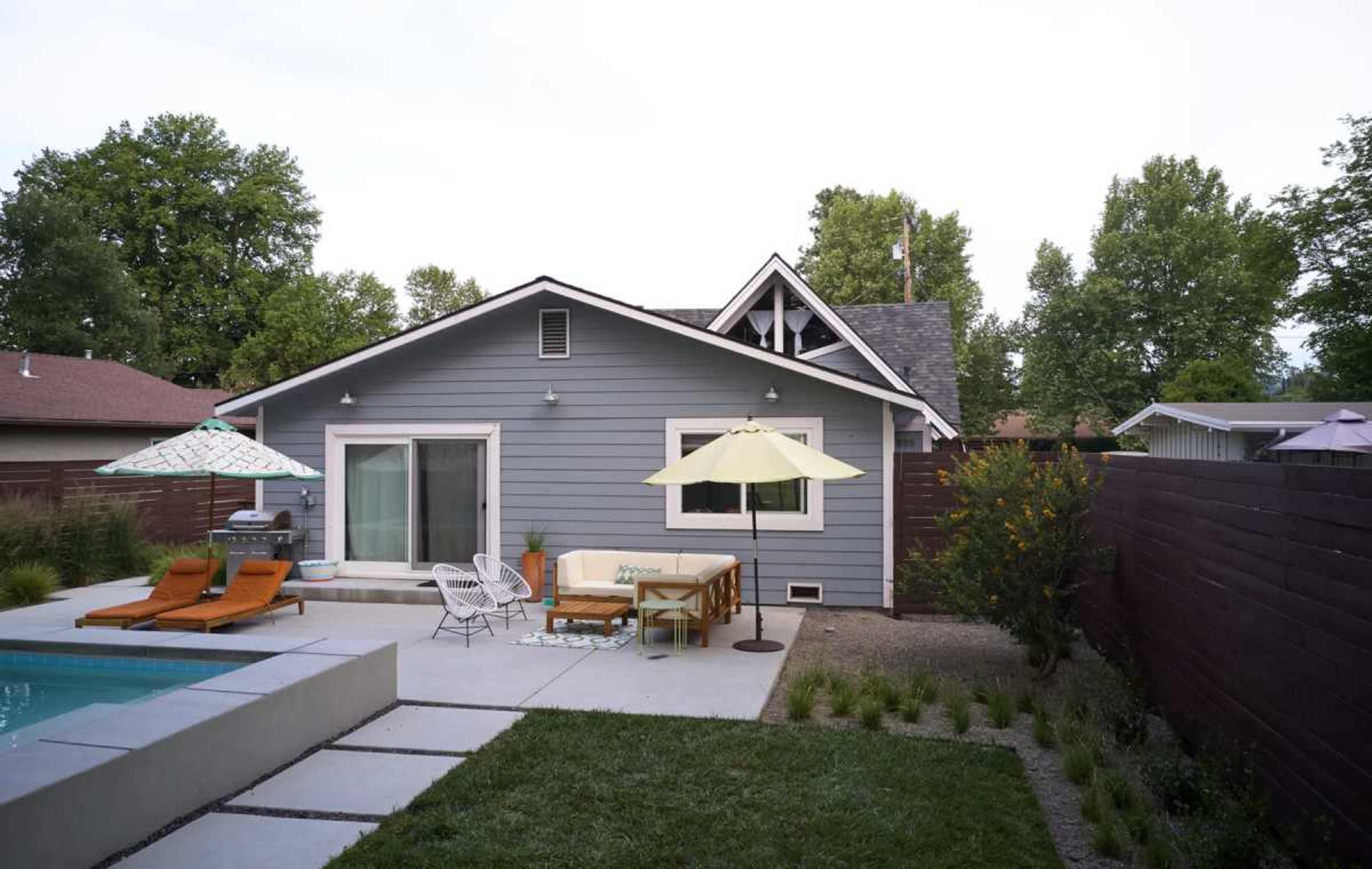A gray house with a small swimming pool and outdoor seating area is surrounded by green trees and a wooden fence.