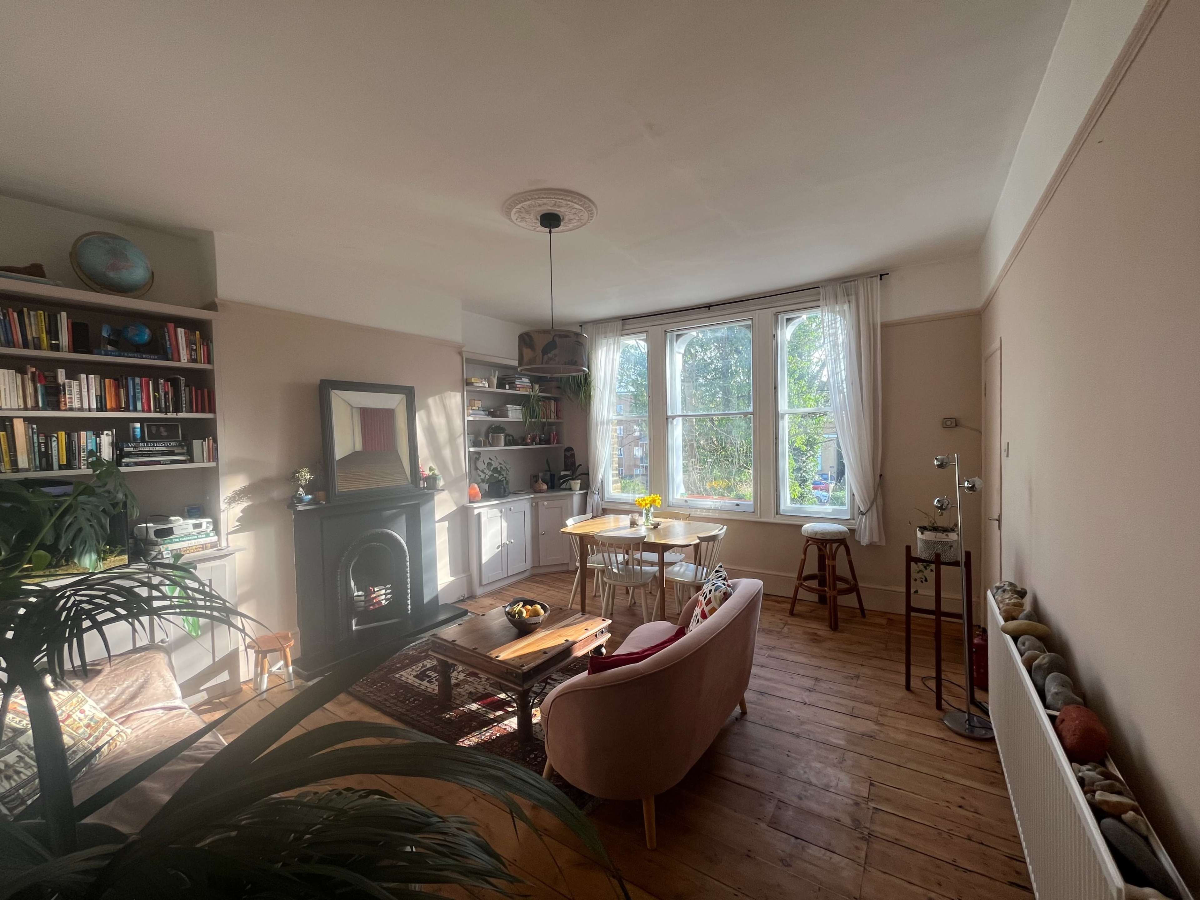 The living room features a pink sofa, a round dining table, bookshelves filled with books, and large windows allowing natural light to illuminate the space.
