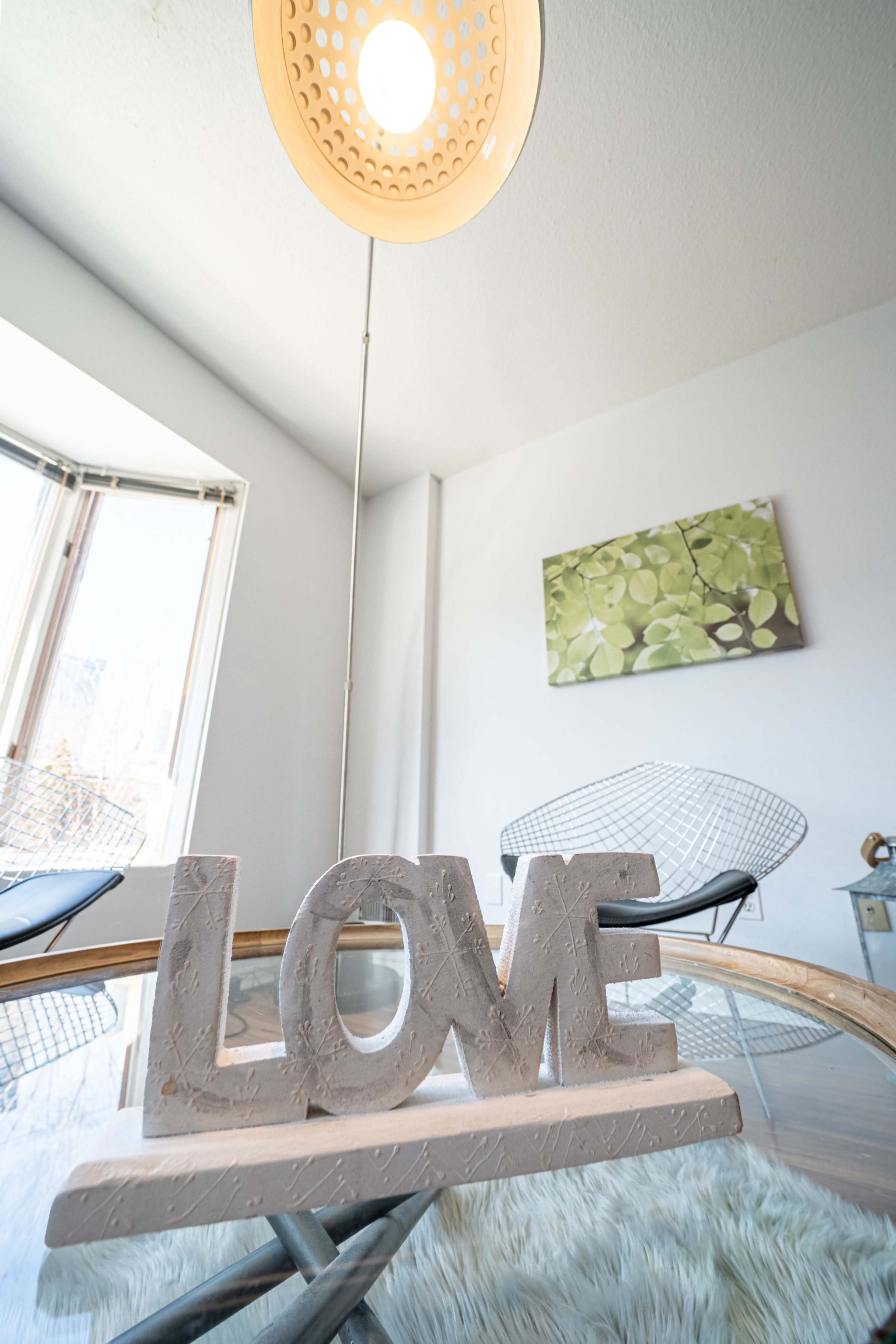 A decorative "LOVE" sign made of concrete is placed on a glass table in a room with a pendant light and a green artwork on the wall.