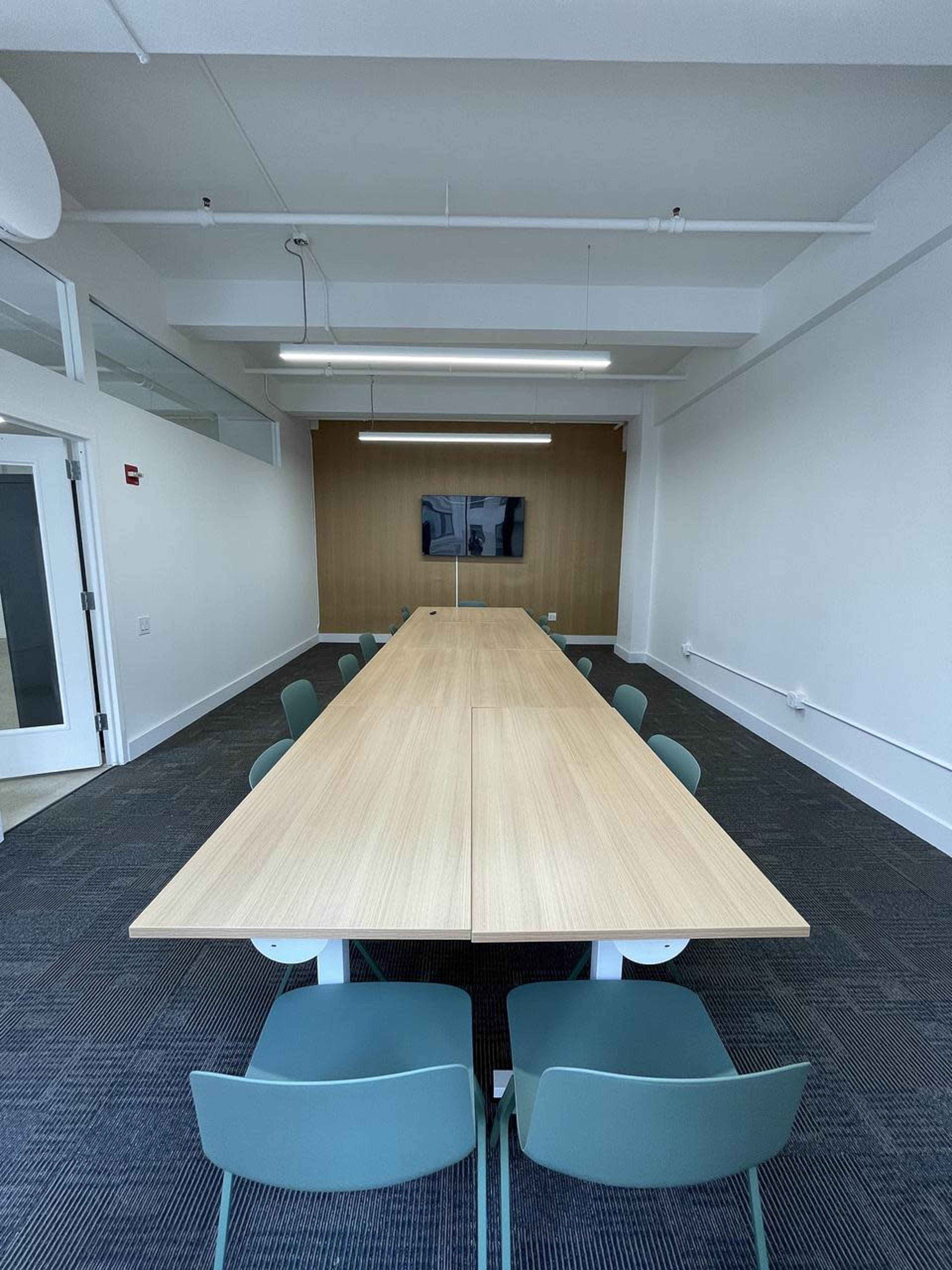 A long, rectangular conference table surrounded by green chairs is positioned in a bright room with a wall-mounted screen in the background.