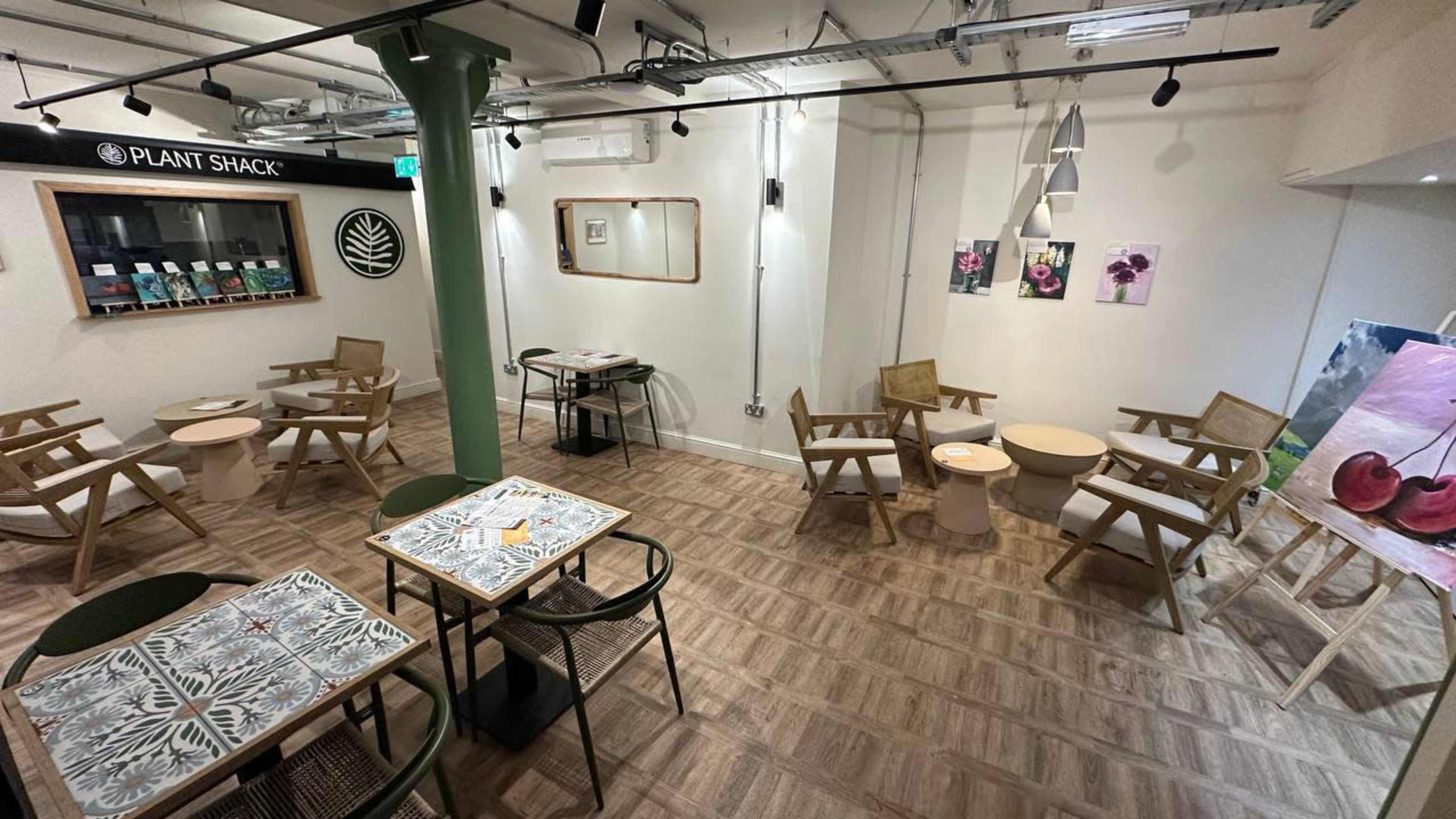 The image shows a modern, minimalist cafe with wooden chairs and tables arranged around a central area, with artwork displayed on the walls.