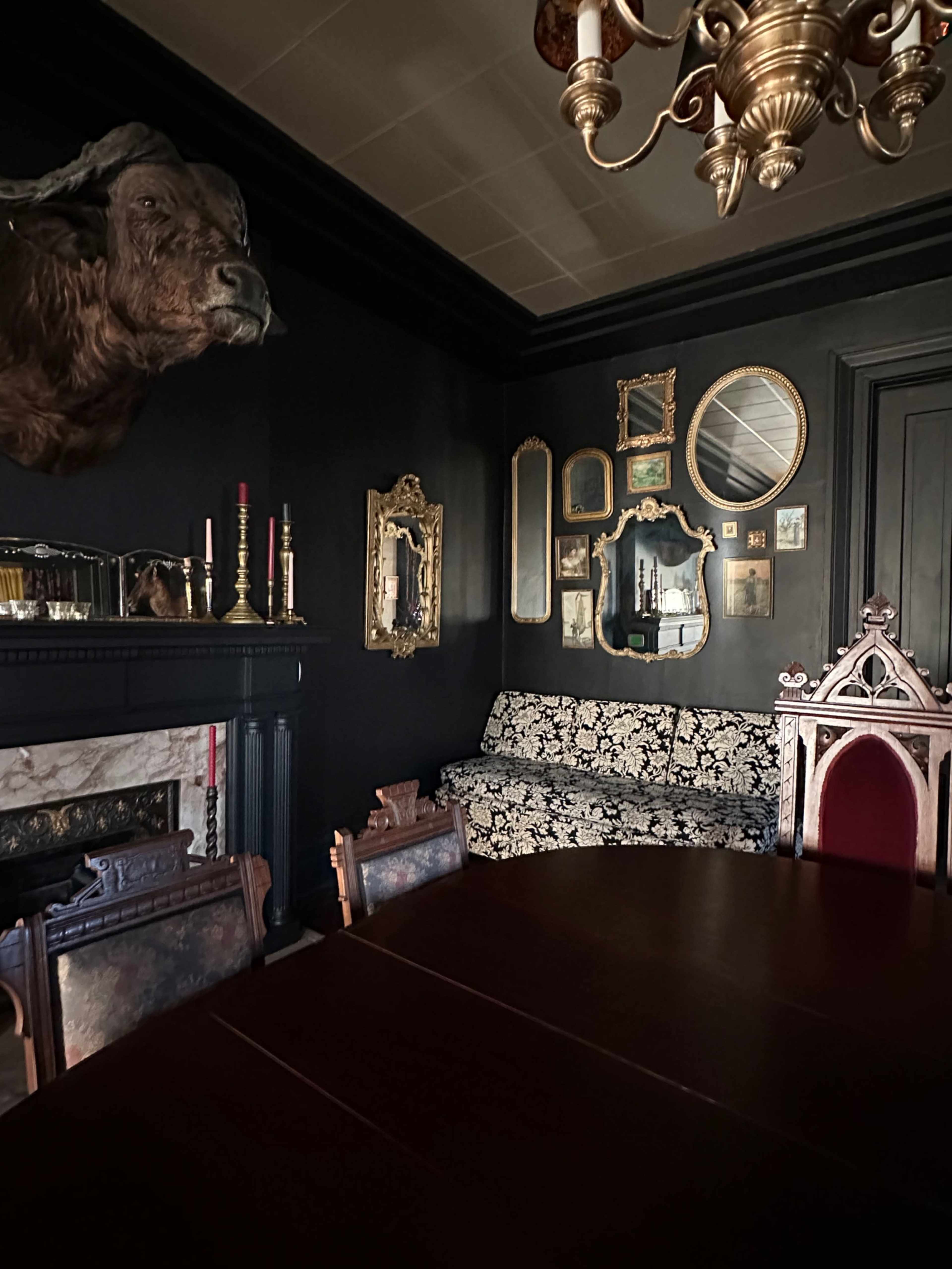 The dimly lit dining room features a dark wall, a mounted animal head above an ornate fireplace, a patterned sofa, and an arrangement of framed mirrors and artwork.