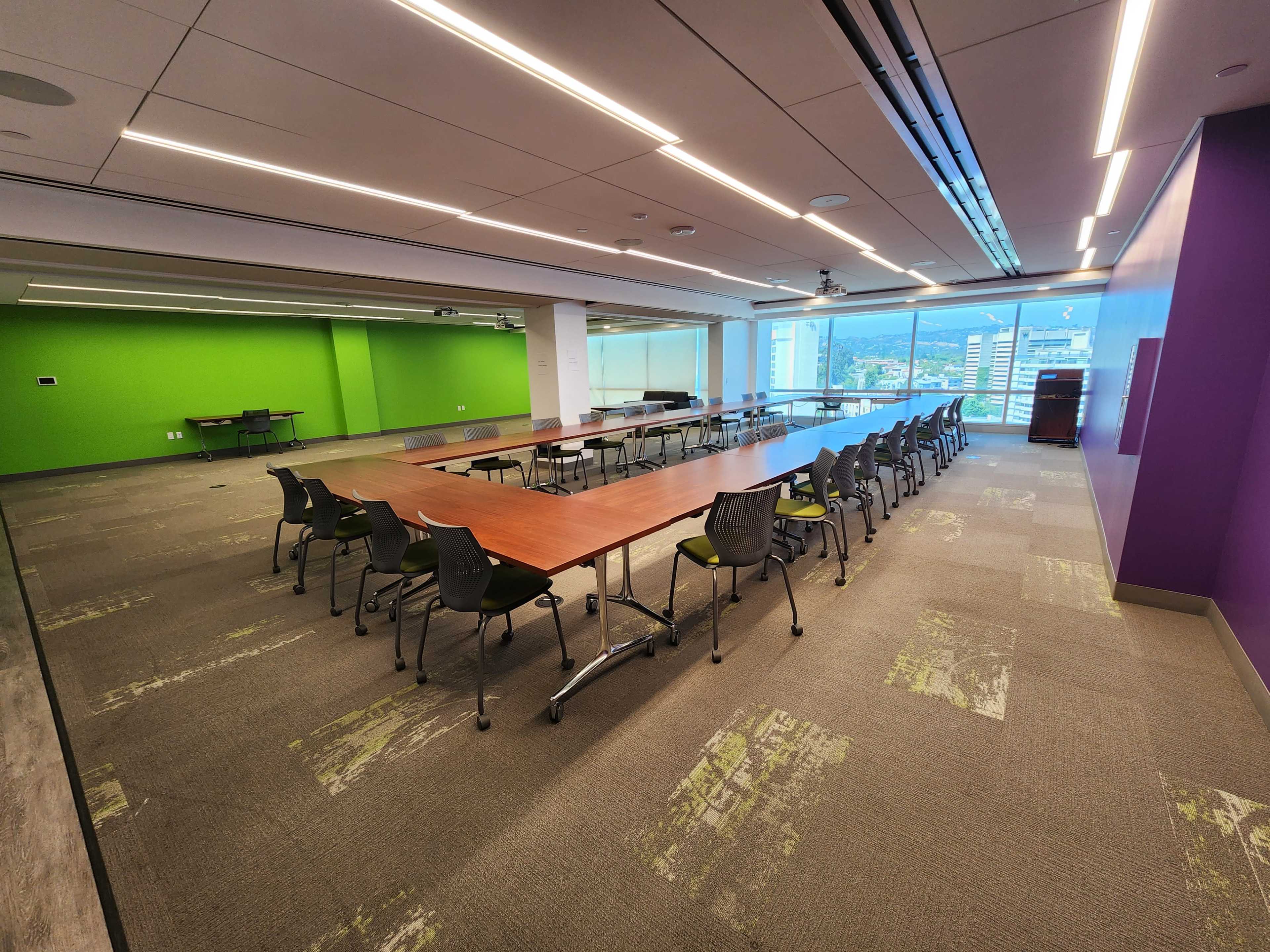 A long conference table is set up in a brightly lit meeting room with green and purple walls and large windows overlooking a cityscape.