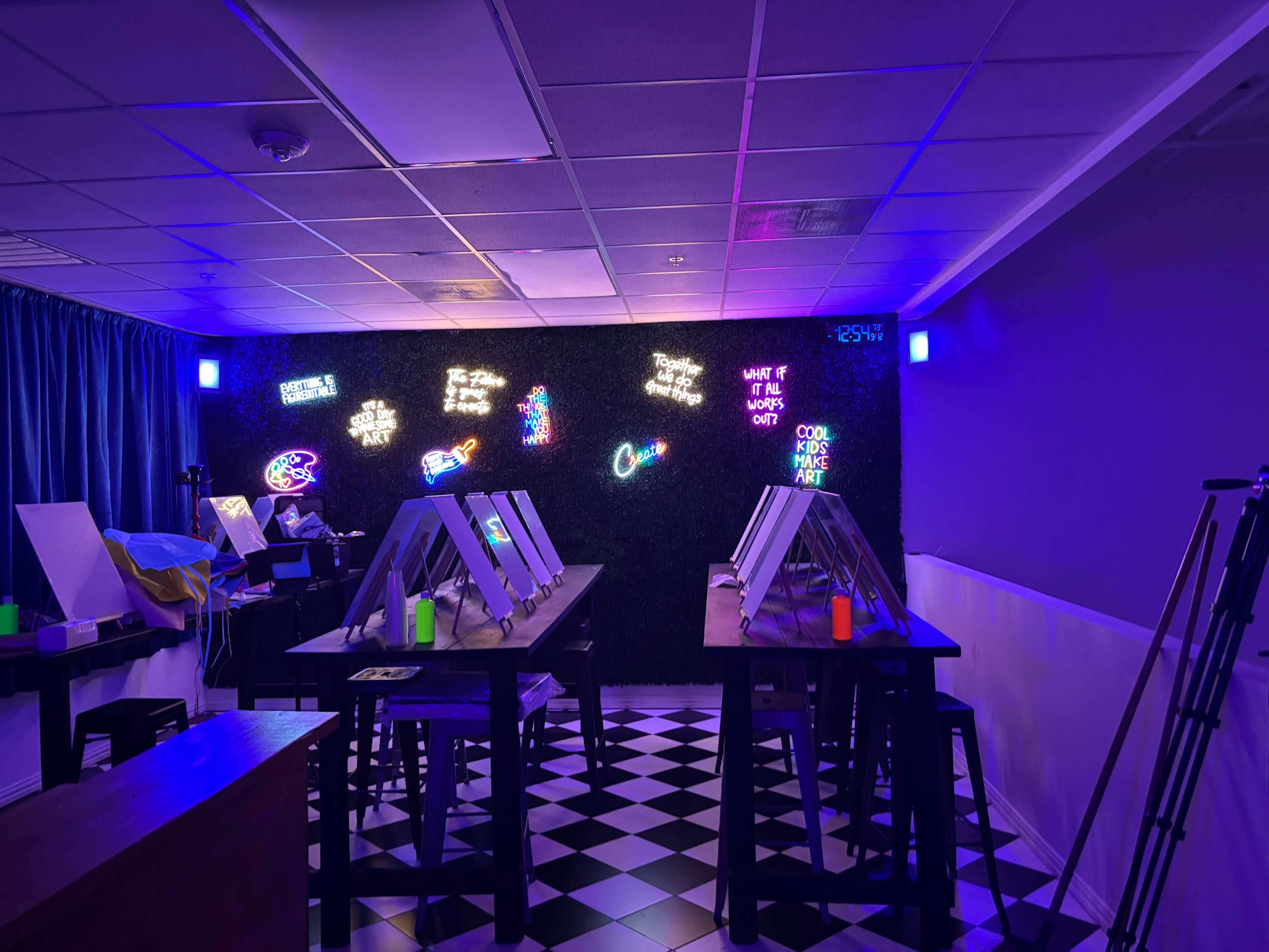 The scene shows a brightly lit art studio with easels, black tables, and neon signs on a dark wall.