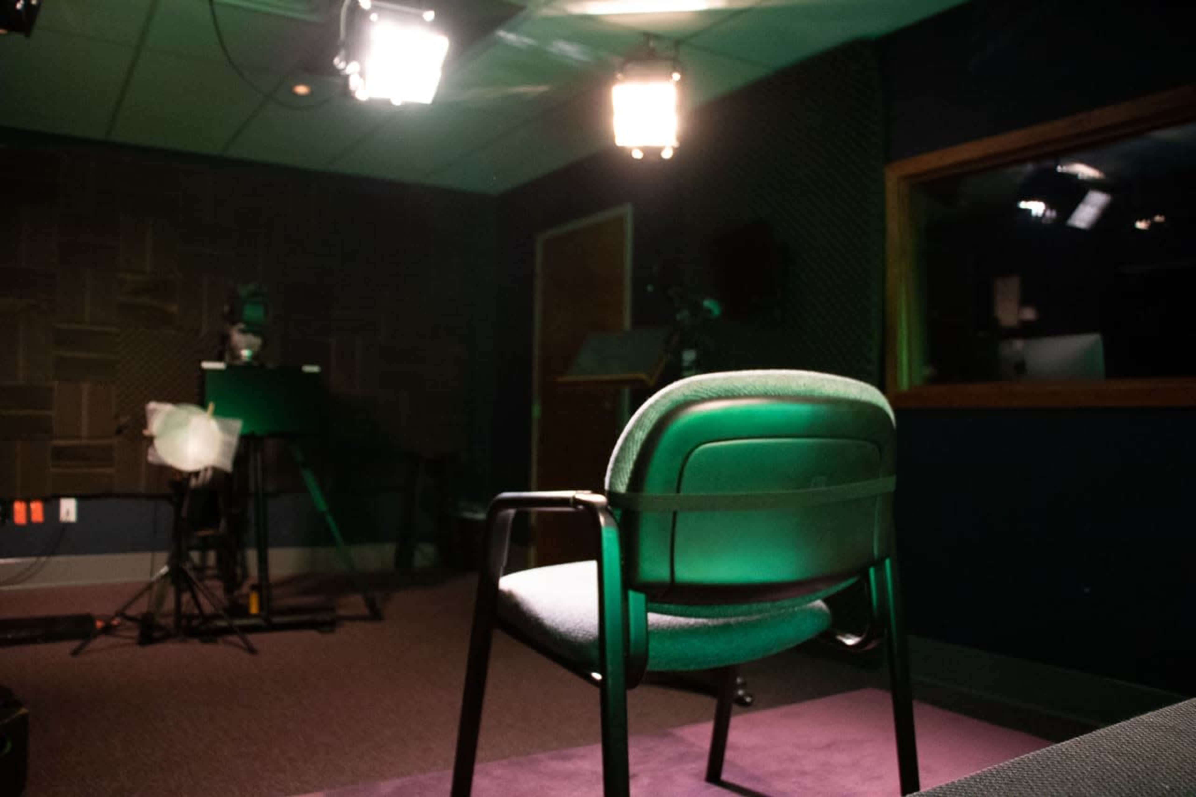 An empty chair positioned in a dimly lit recording studio with various equipment visible in the background.
