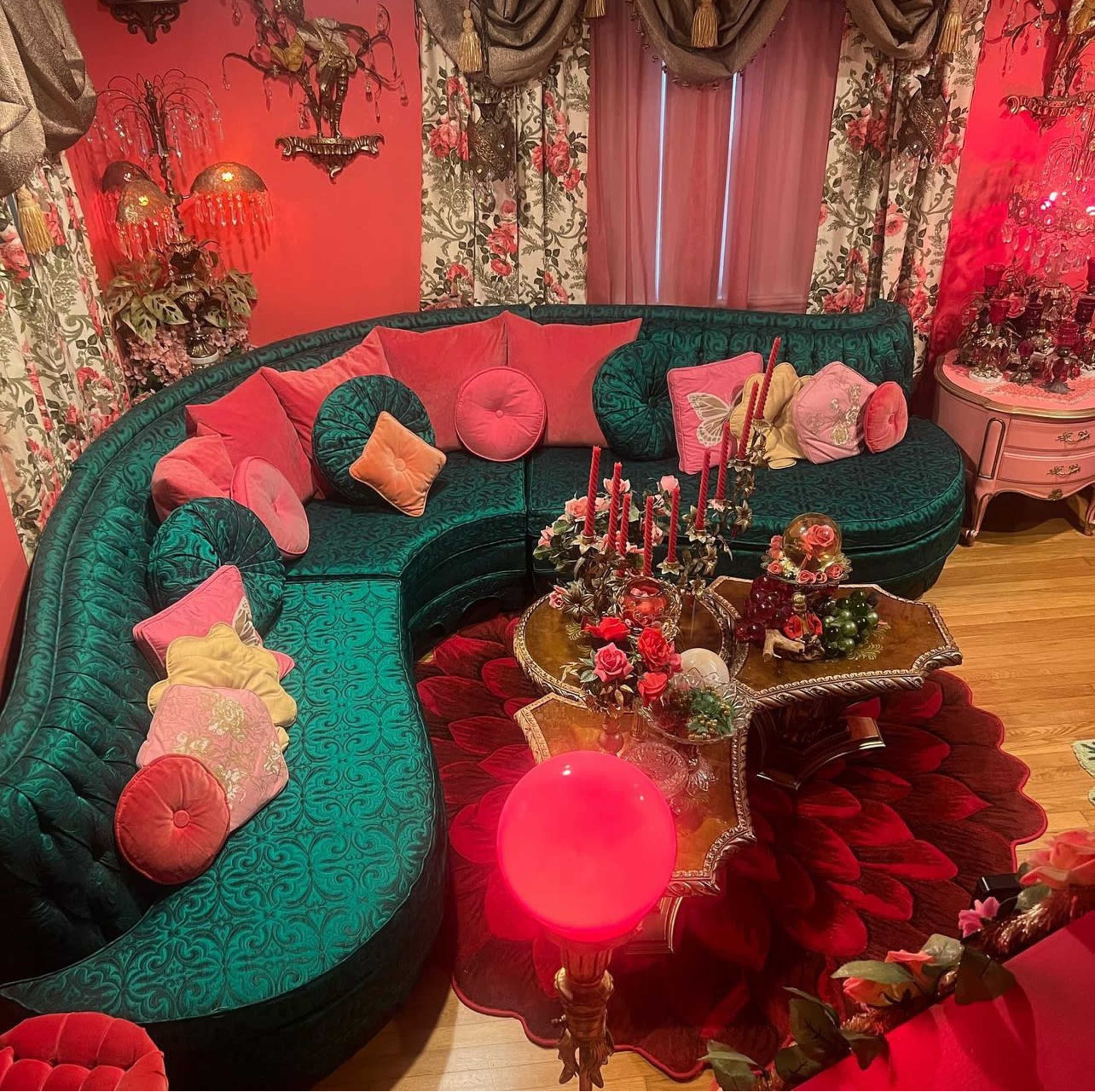The image shows a brightly decorated living room featuring a large green sectional sofa adorned with various cushions, a floral-themed coffee table, and decorative candles, all set against pink walls.