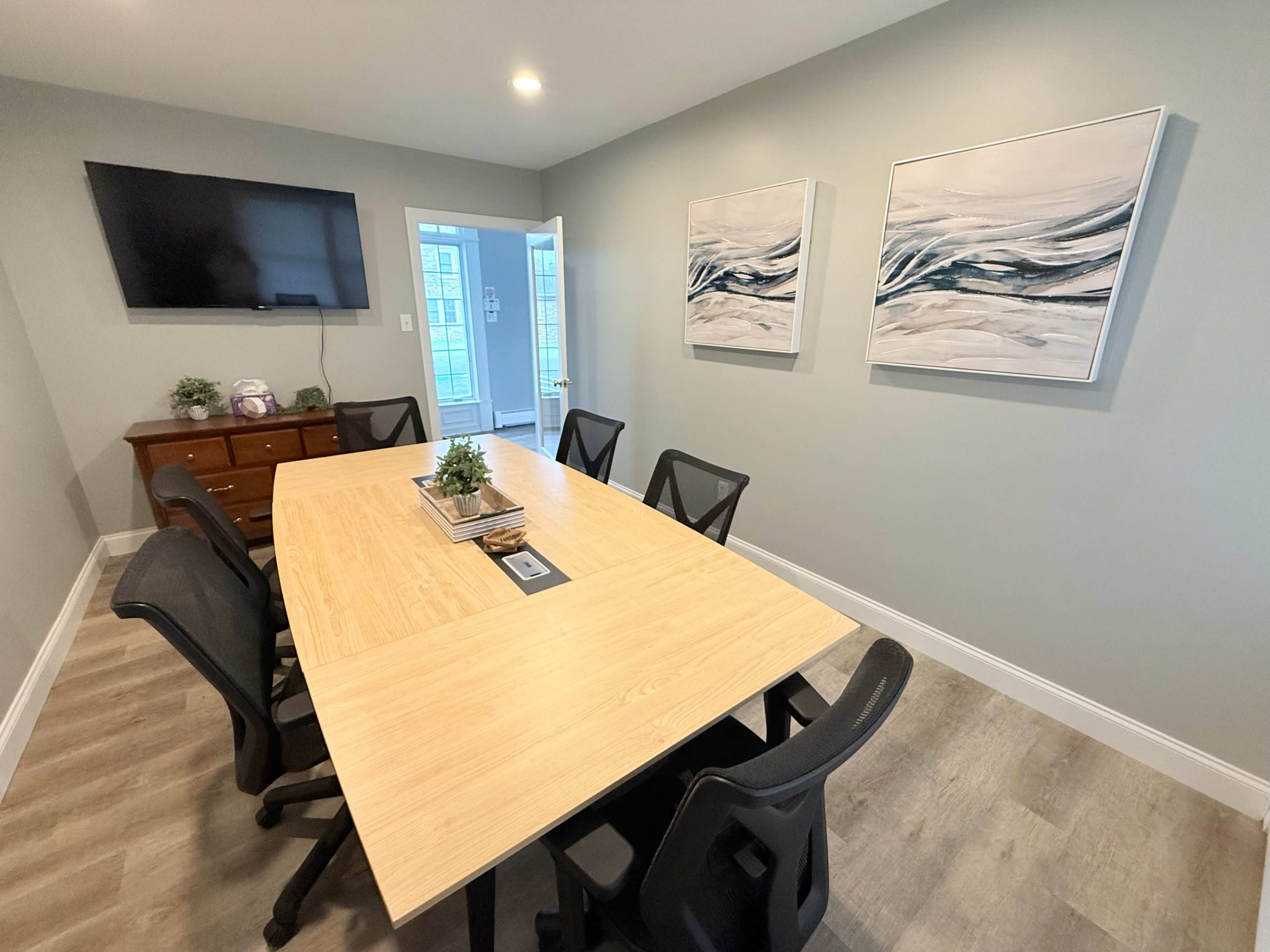A conference room features a large wooden table surrounded by black chairs, with two abstract paintings on the wall and a TV mounted in the corner.