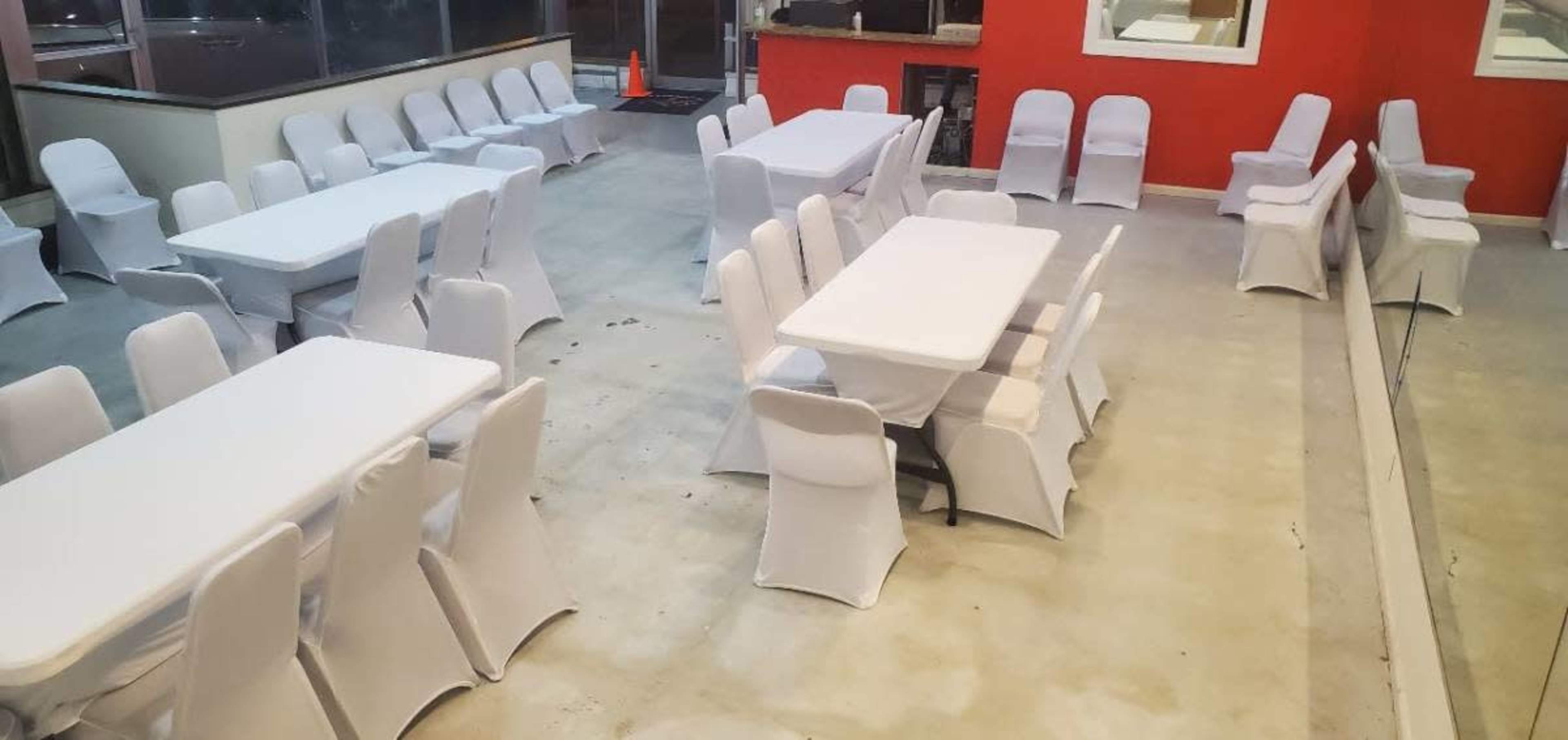 The image shows a spacious indoor venue arranged with multiple white-covered tables and chairs facing one another, along with a red accent wall and large windows.