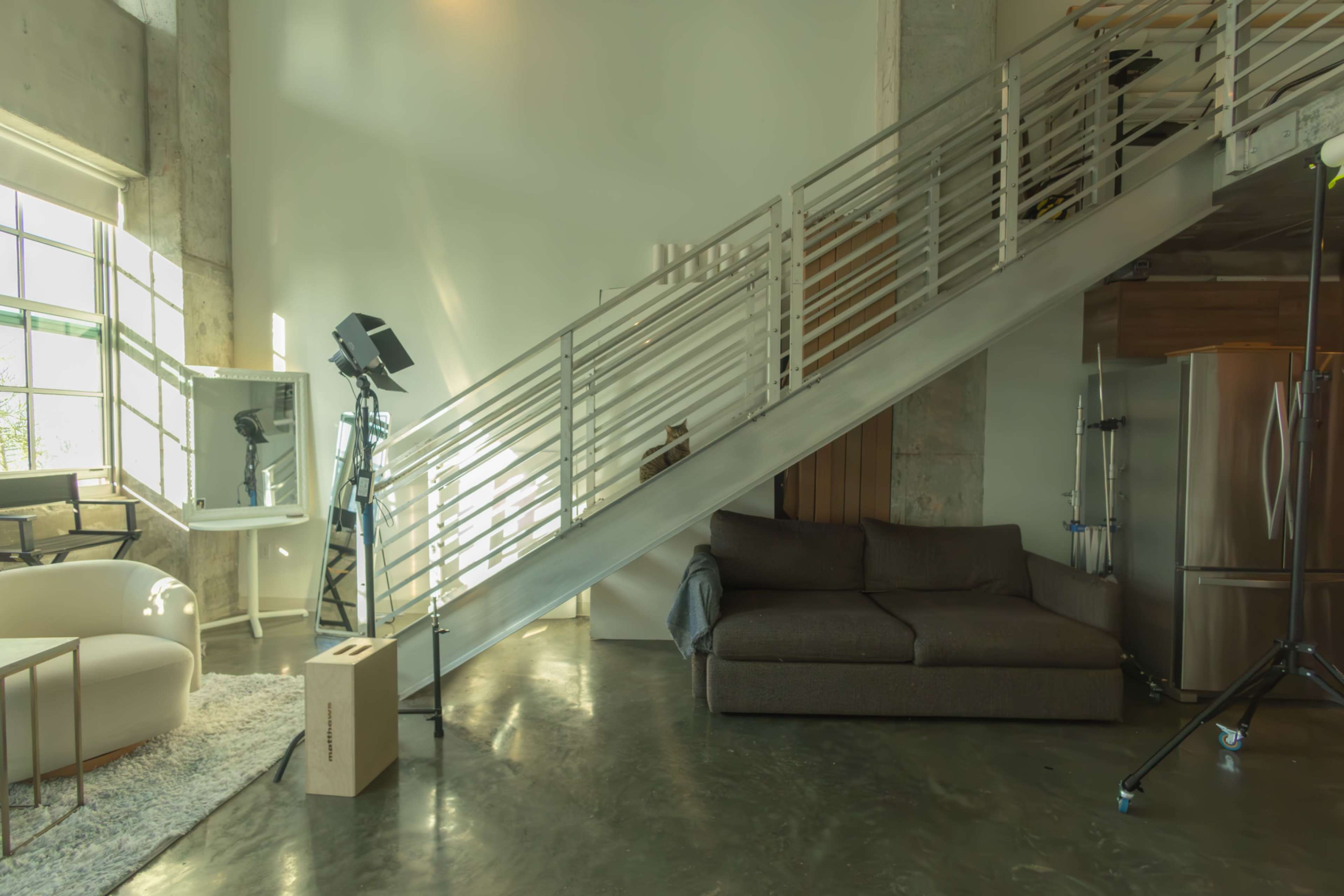 The image shows a modern interior space featuring a staircase, a sofa, a mirror, and lighting equipment, with large windows allowing natural light to enter.
