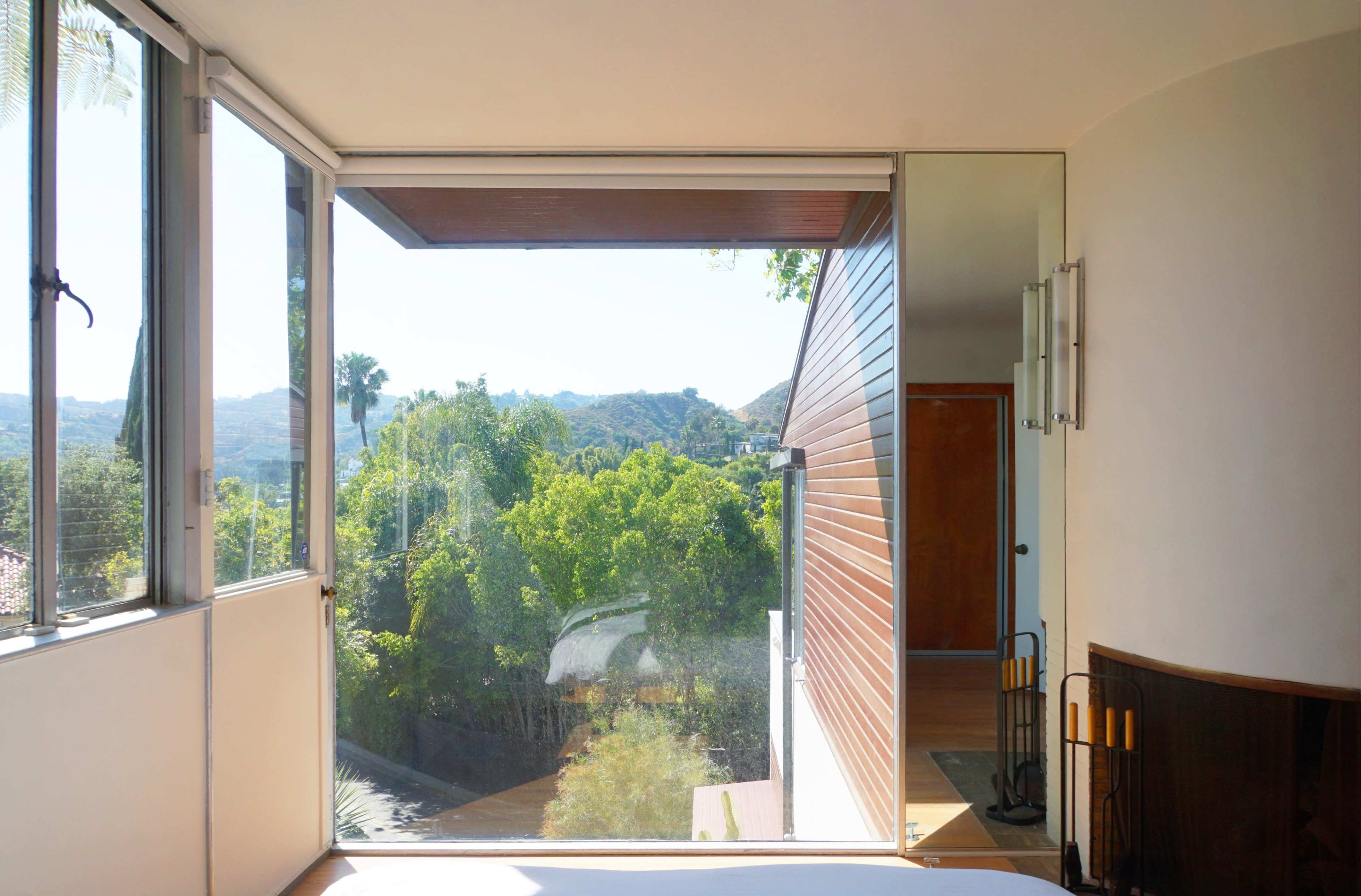 A modern bedroom features large glass windows overlooking a landscaped green area and distant hills.