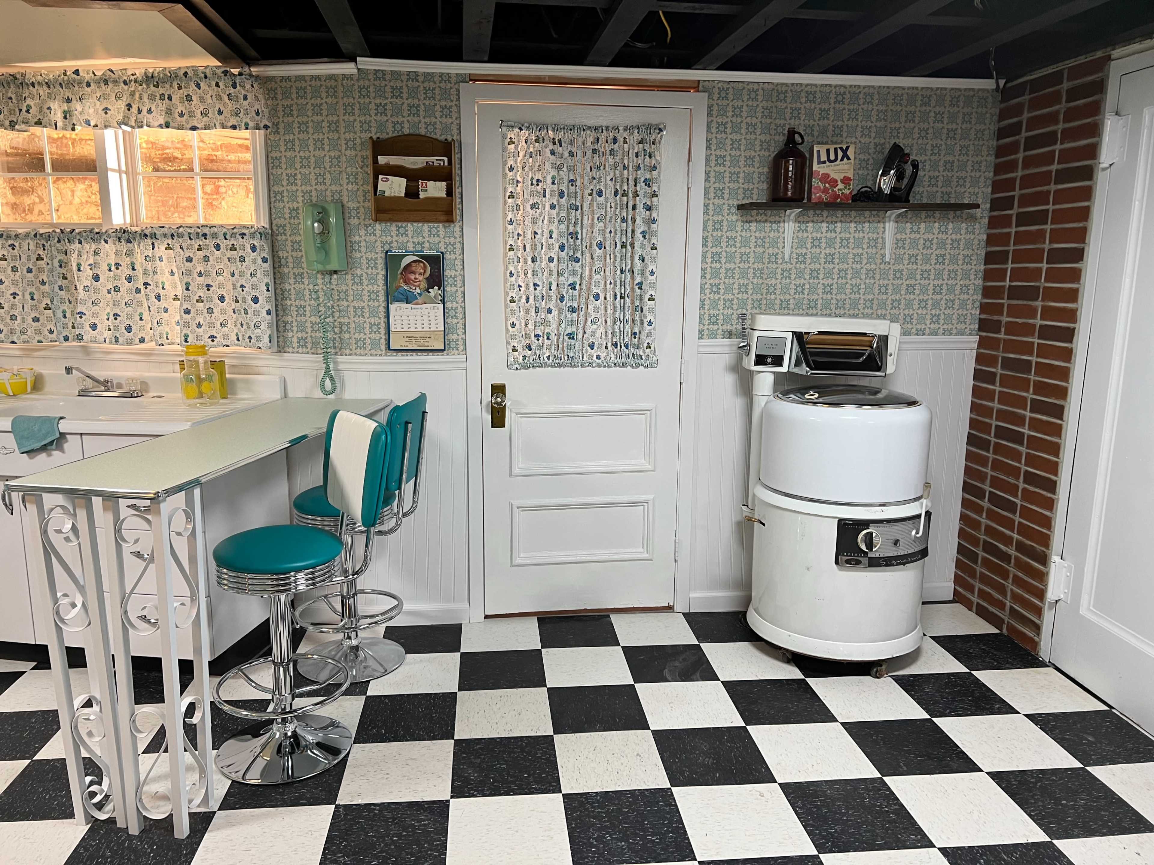 A vintage kitchen with a checkered black and white floor, a white door, a retro washing machine, and teal bar stools at a counter.