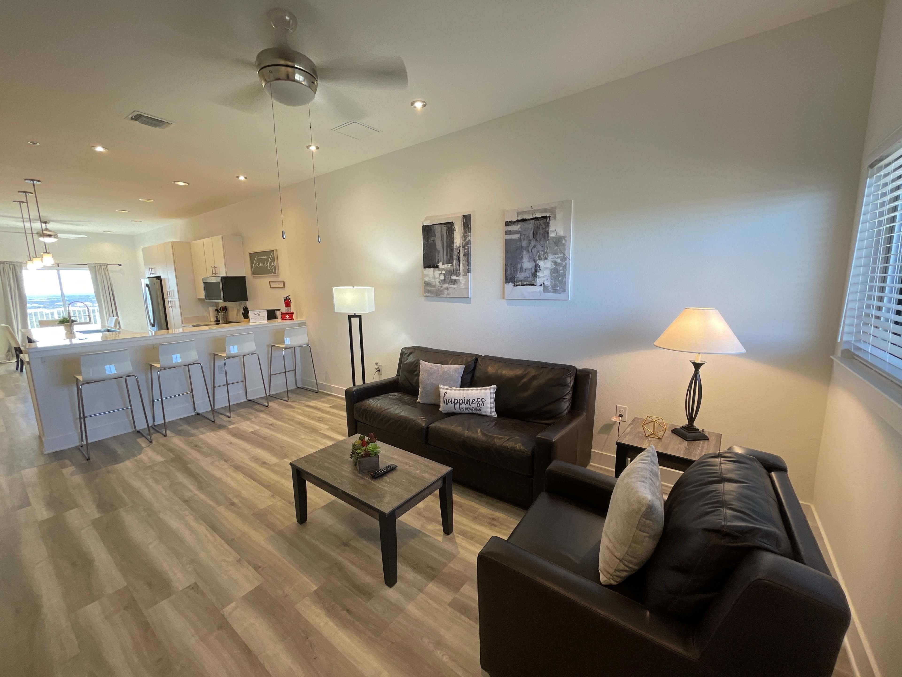 A modern living room with two black leather sofas, a coffee table, and a dining area and kitchen visible in the background.