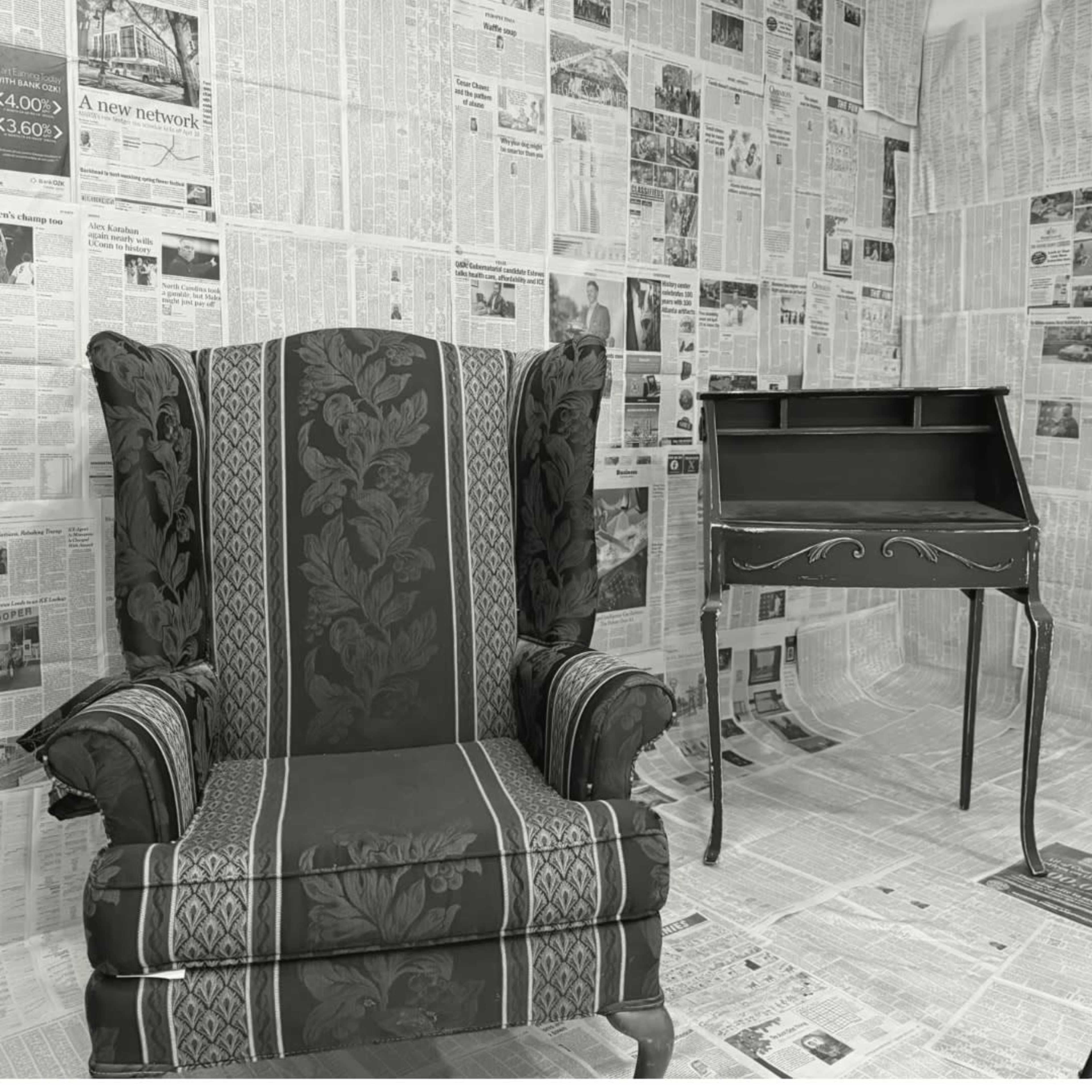 A vintage wingback chair and a small writing desk are positioned in a room with walls and floor covered in newspapers.
