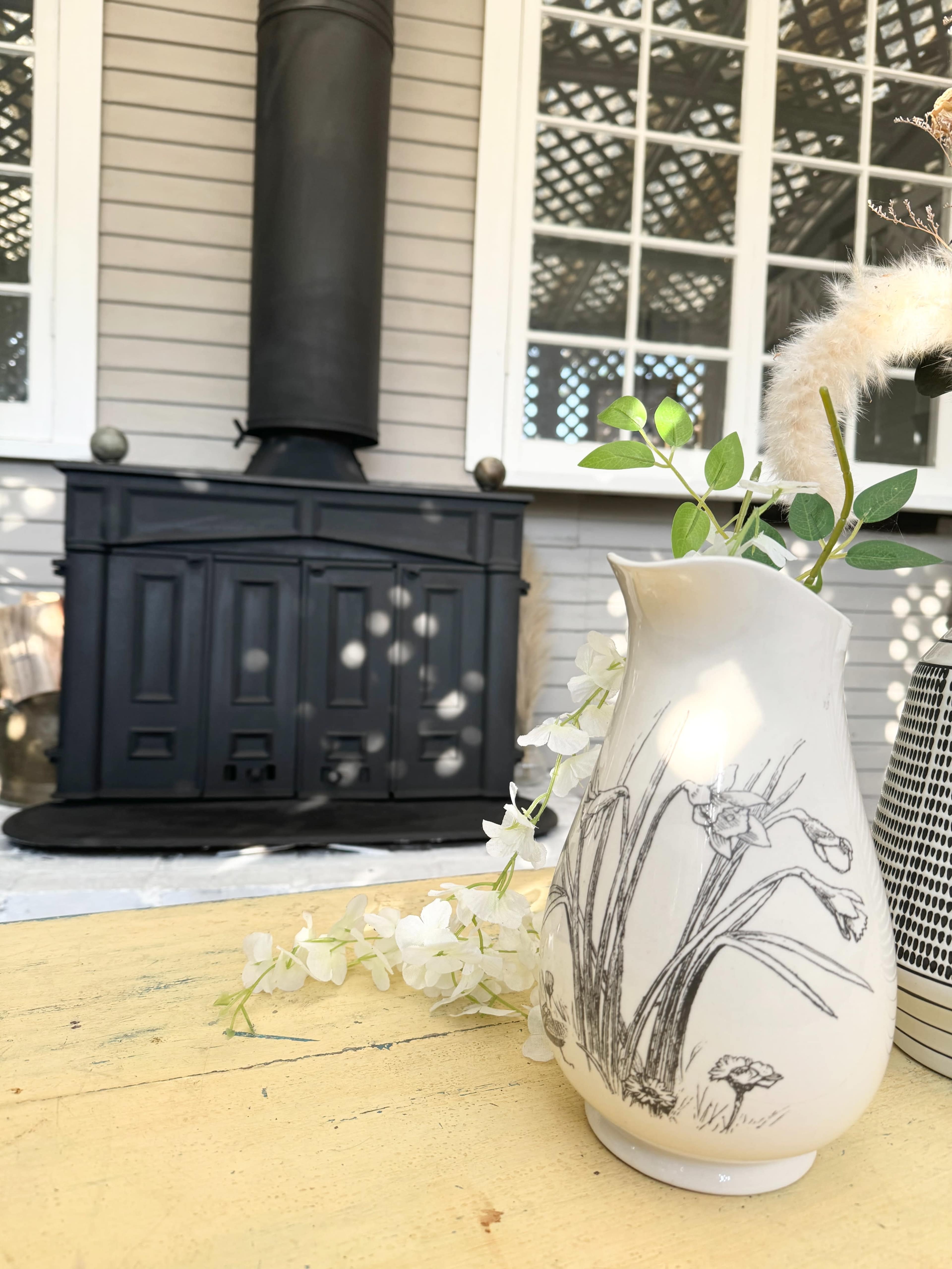 A white vase with a floral design sits on a yellow table in front of a black wood stove and a window with a lattice pattern.