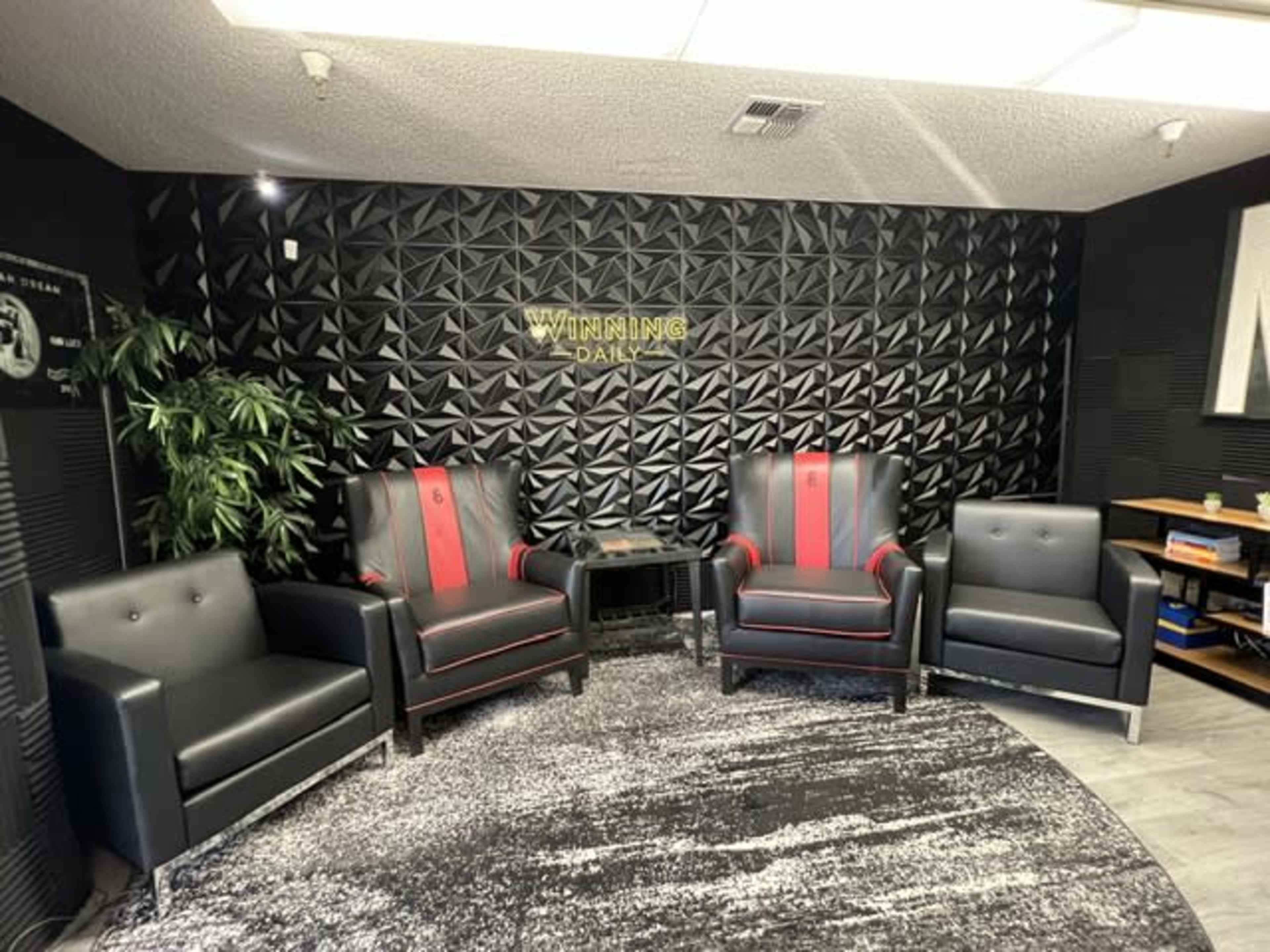 The image shows a modern meeting space featuring four black chairs with red accents arranged around a round rug, against a textured wall with the word "WINNING" displayed prominently.