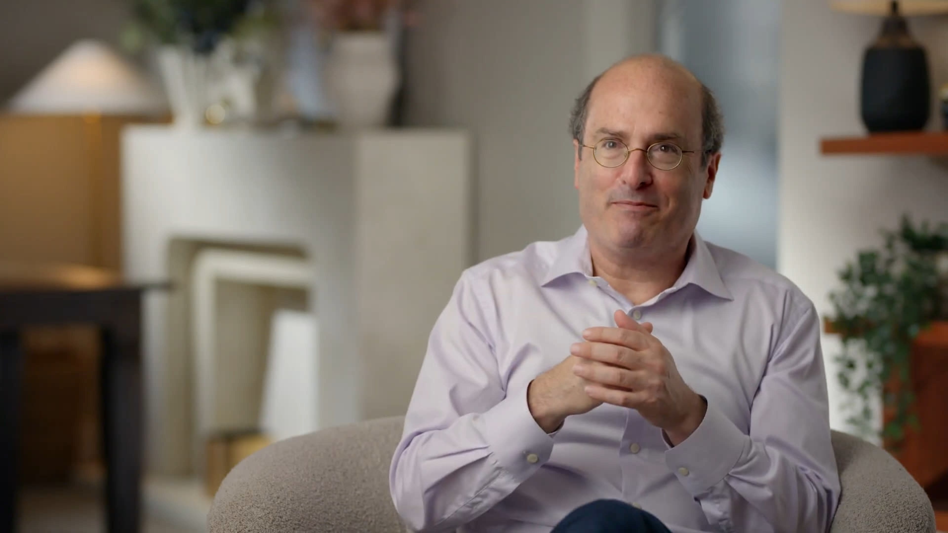 A man with glasses sits in a light-colored chair, clasping his hands together, in a softly lit room with a cozy atmosphere.