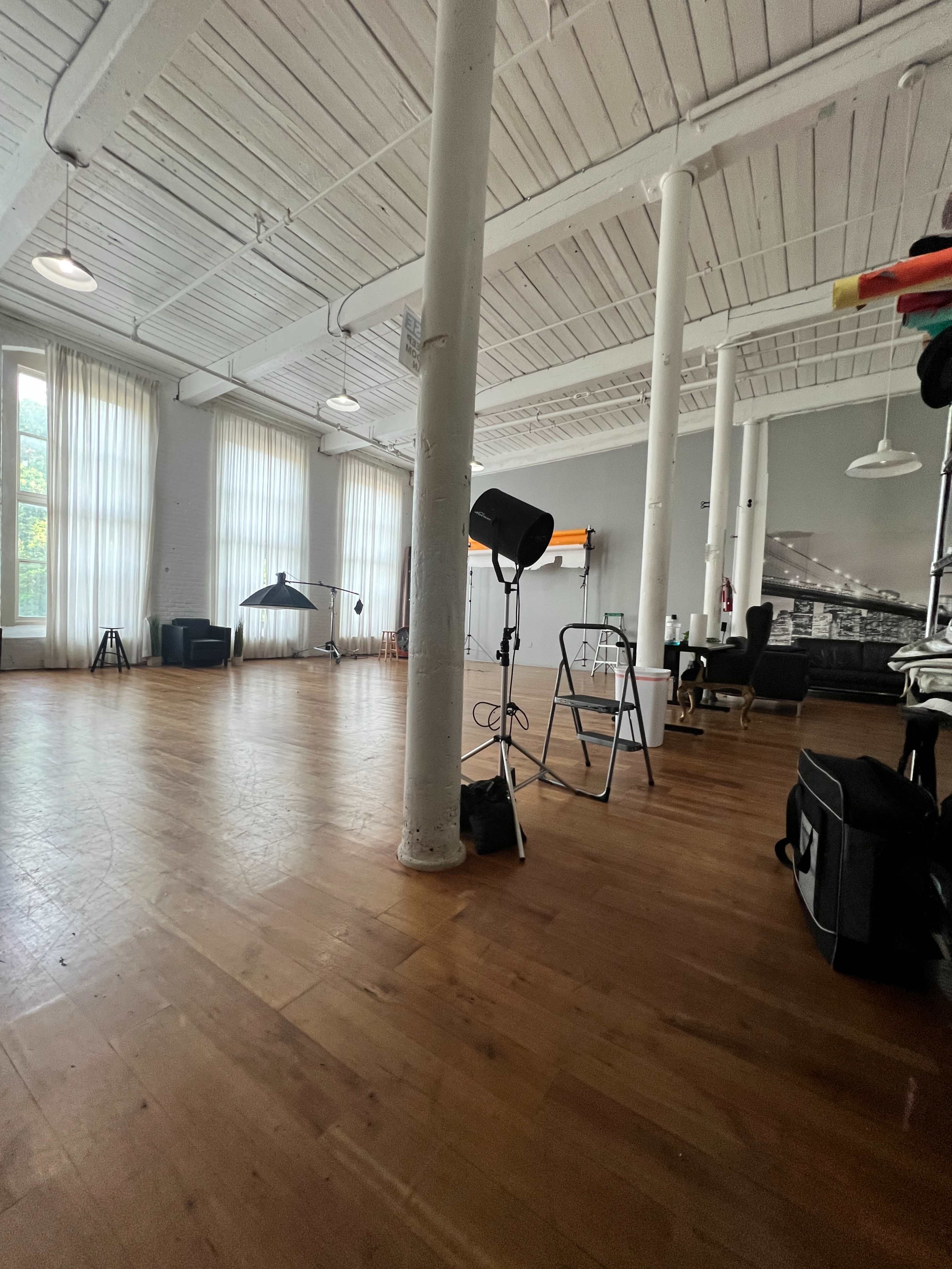 The image shows a spacious, well-lit studio with wooden flooring, large windows, and various equipment including a light stand and a ladder.