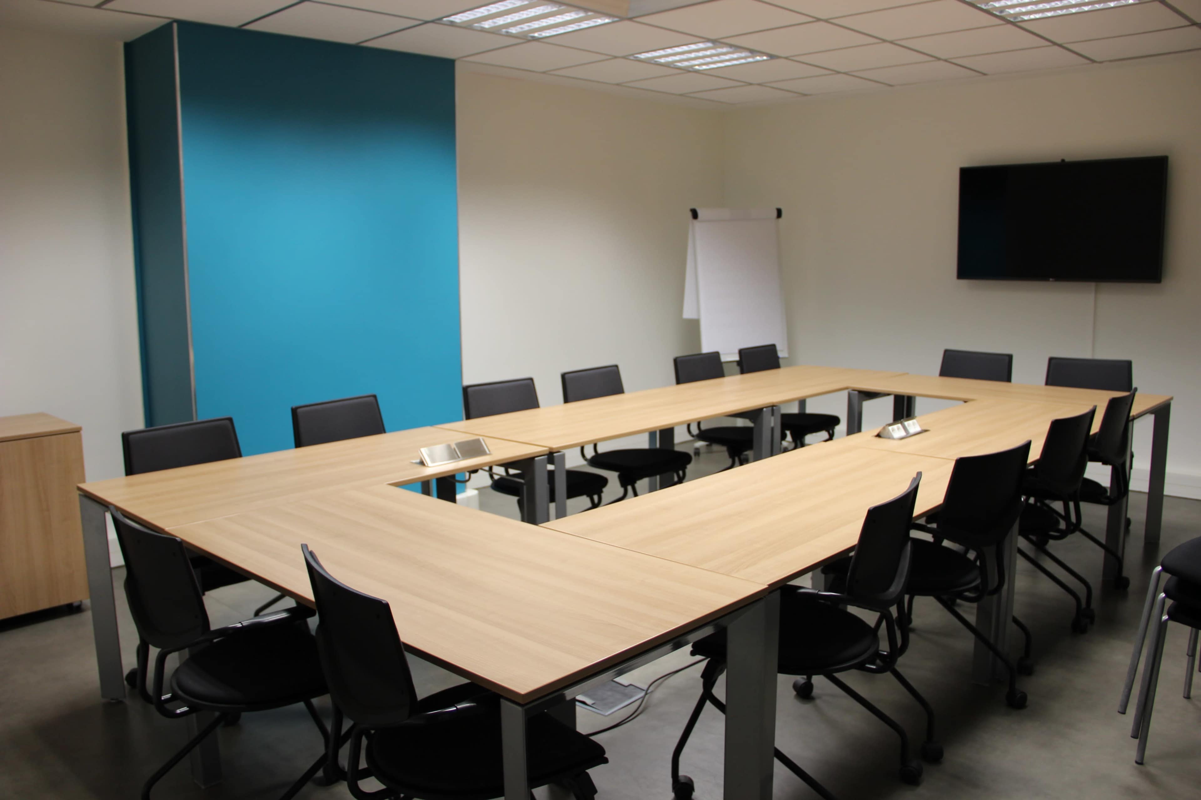 A conference room features a large rectangular table surrounded by black chairs, with a blue accent wall and a television mounted on the opposite wall.