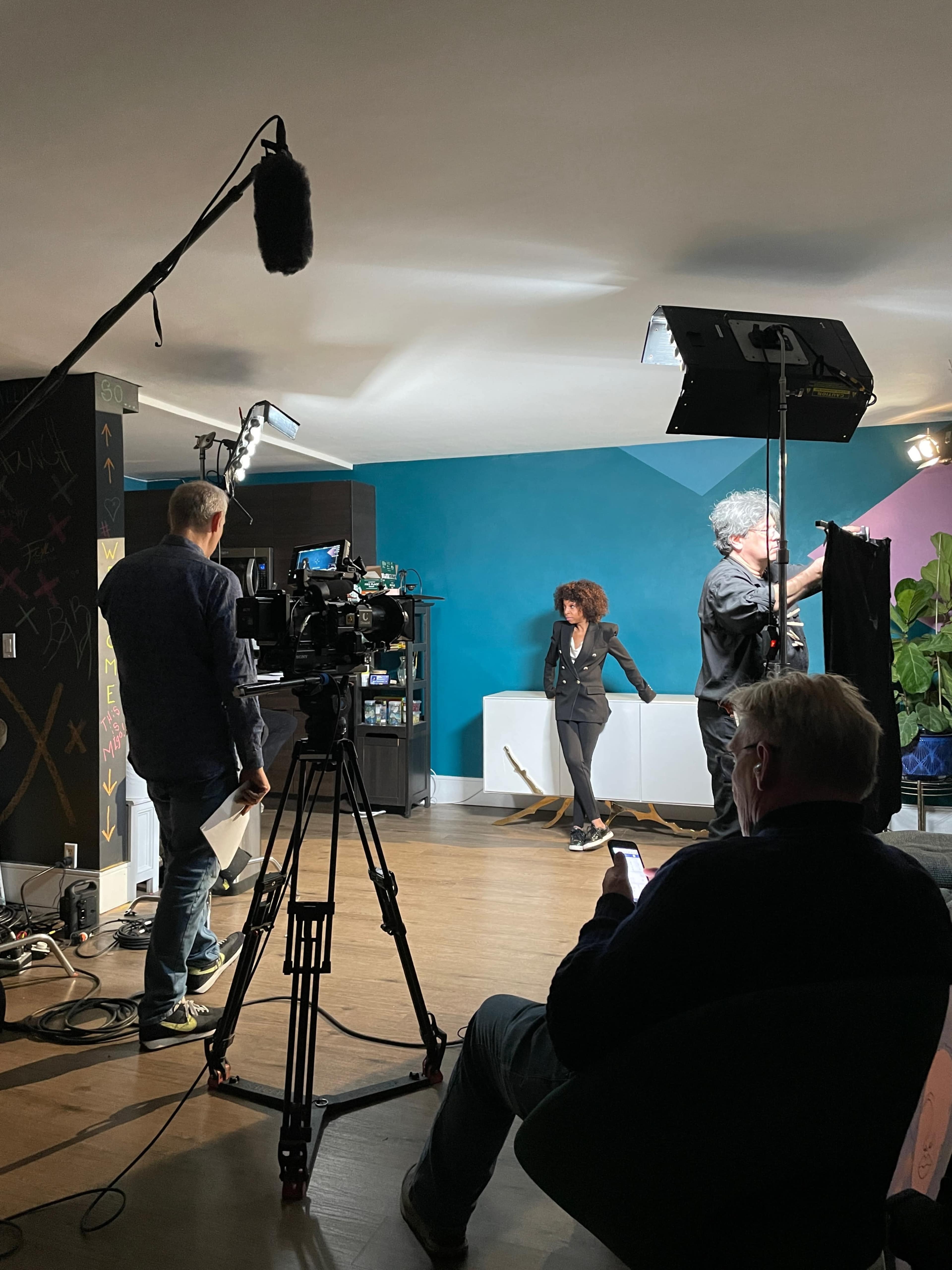 A film crew sets up a shot in a colorful studio with a woman standing in front of a backdrop while a man adjusts lighting equipment.