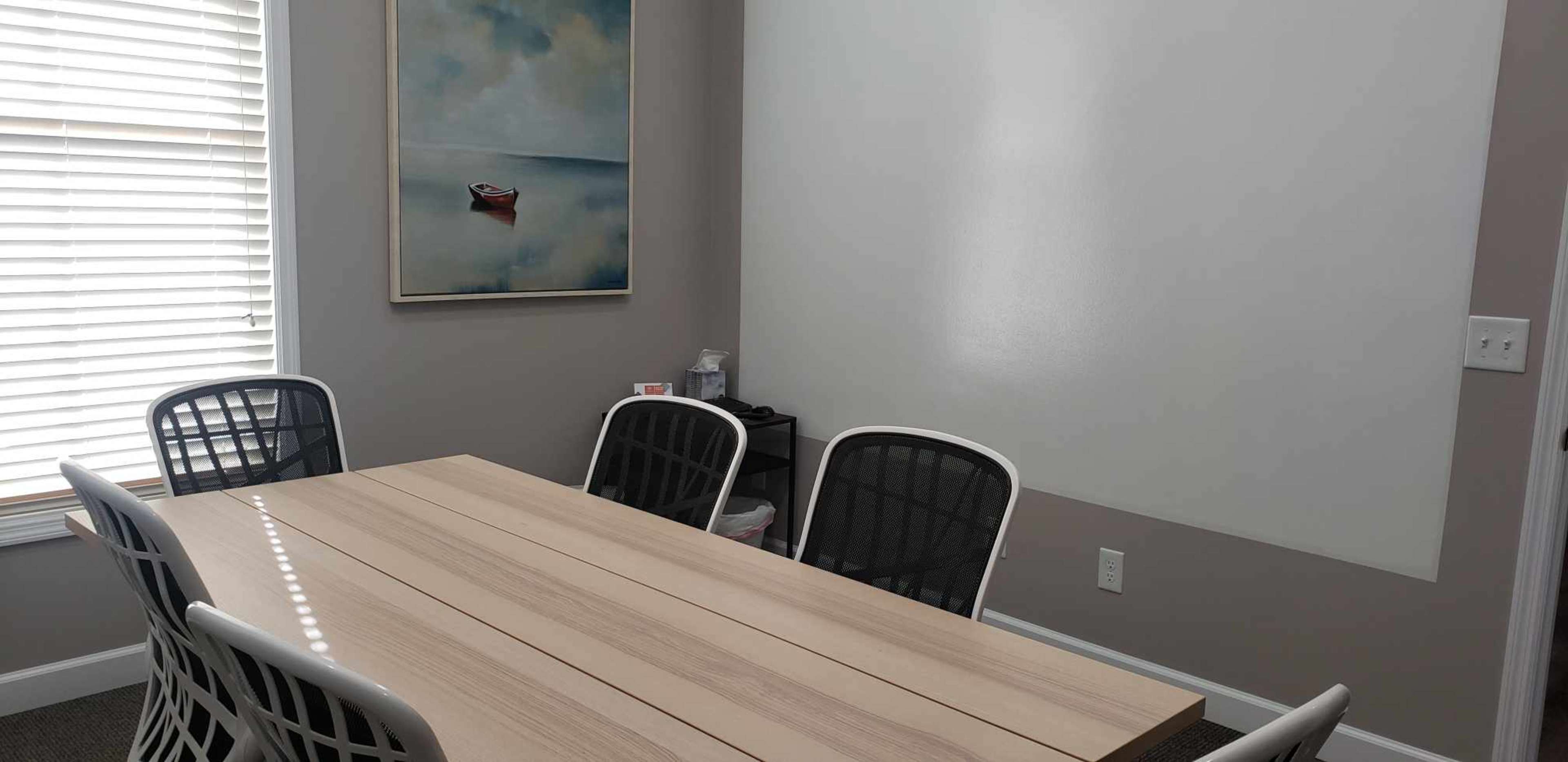 A meeting room features a wooden table surrounded by four chairs, with a painting of a boat on the wall and windows with horizontal blinds.