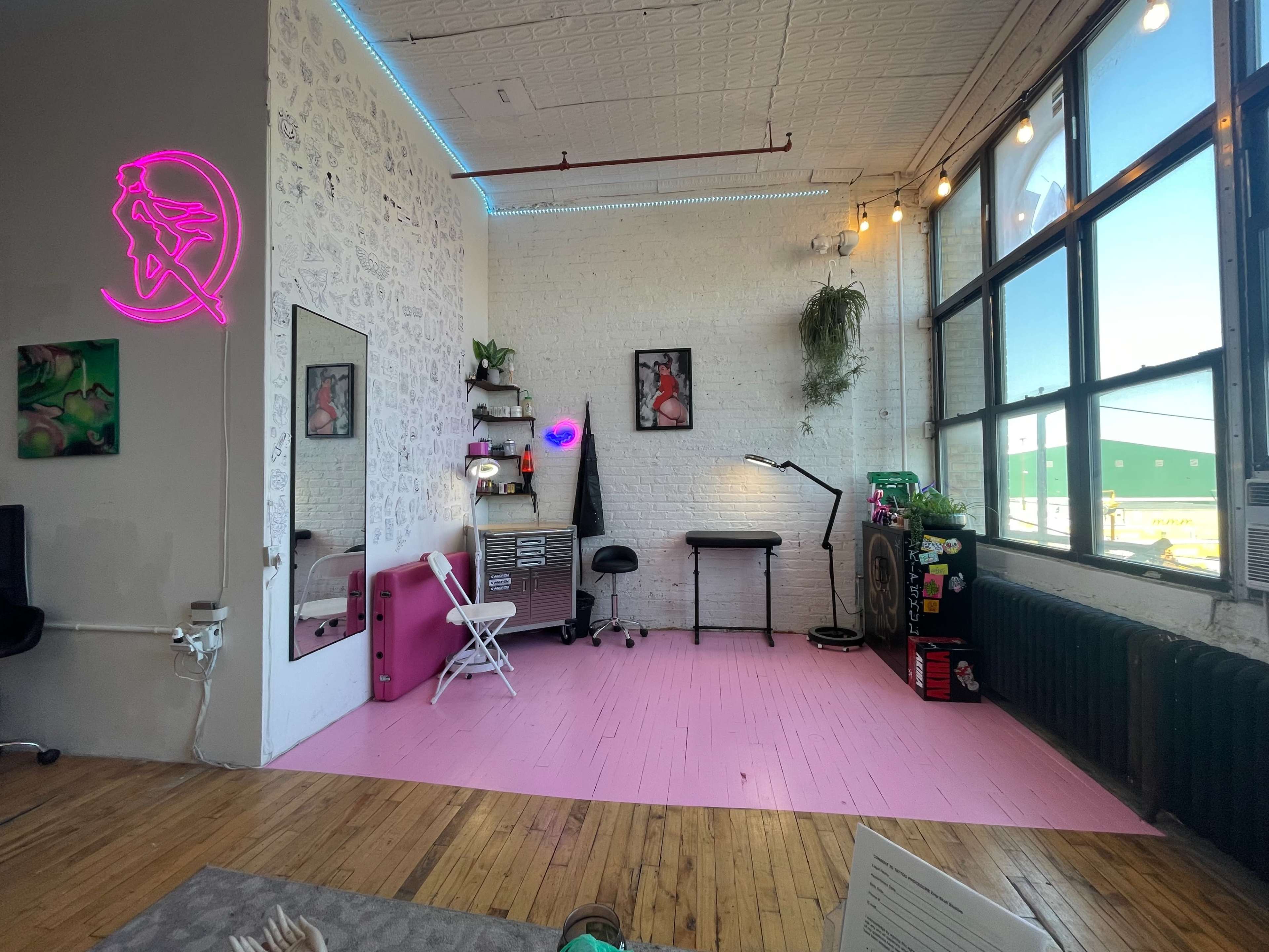 The image shows a bright, modern workspace featuring a pink floor, a work table, a chair, and large windows that allow natural light to enter the room.