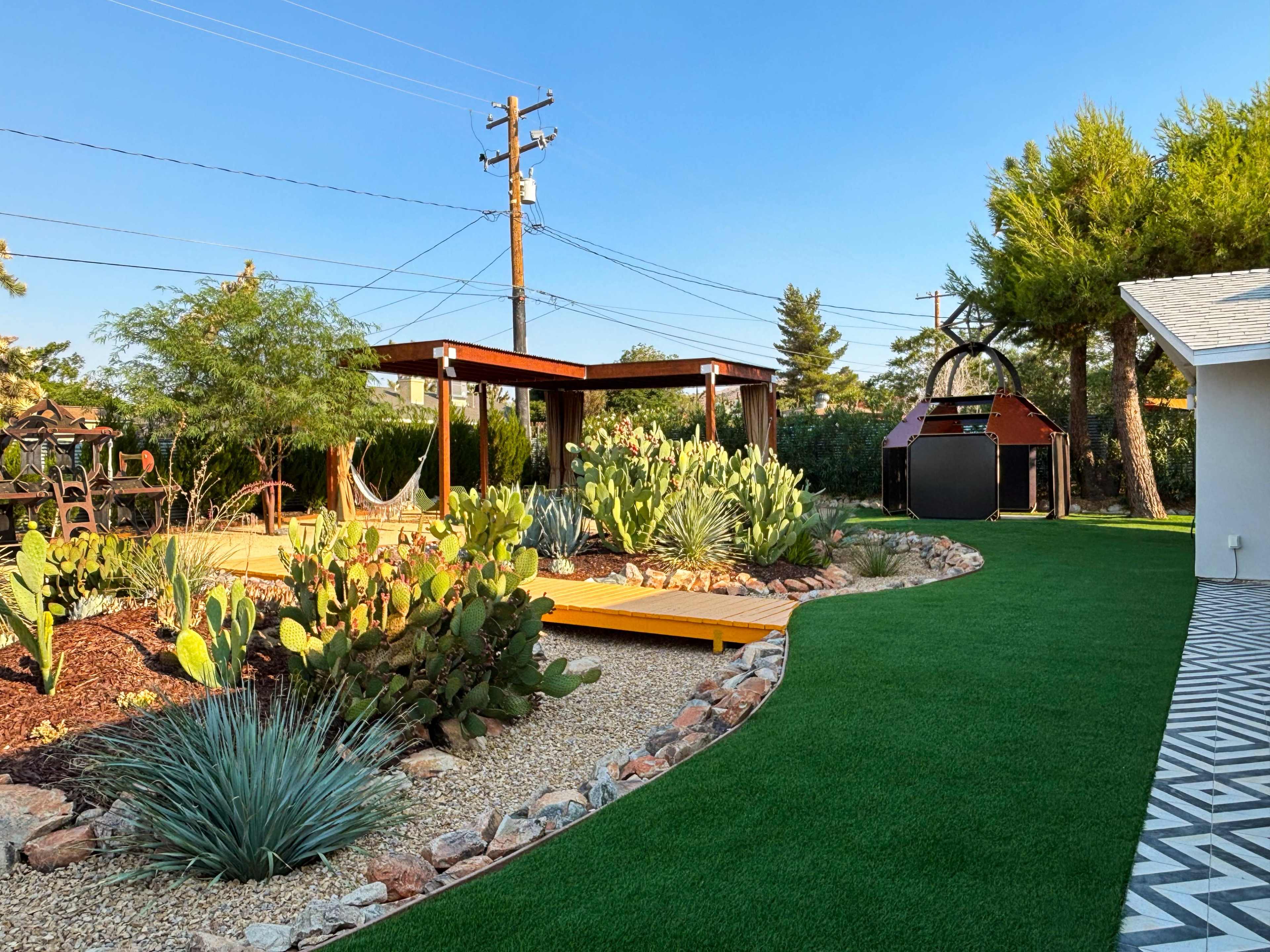 The image shows a landscaped backyard featuring a wooden pergola, a hammock, various cacti, artificial grass, and a section of patterned paving.