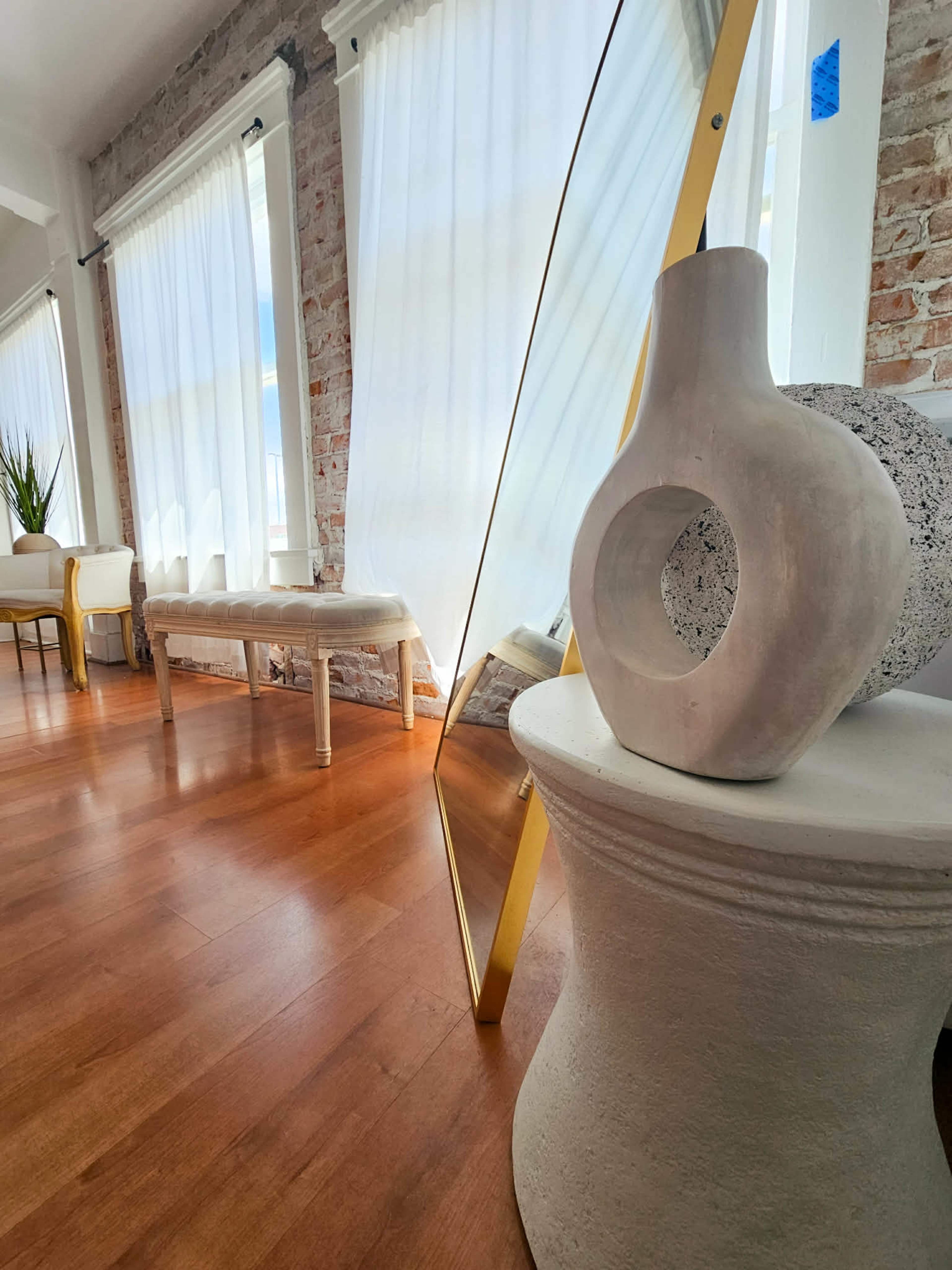 The image shows a well-lit interior space featuring large windows with sheer curtains, a decorative vase, and a mirror reflecting the room's elements, all set on wooden flooring.