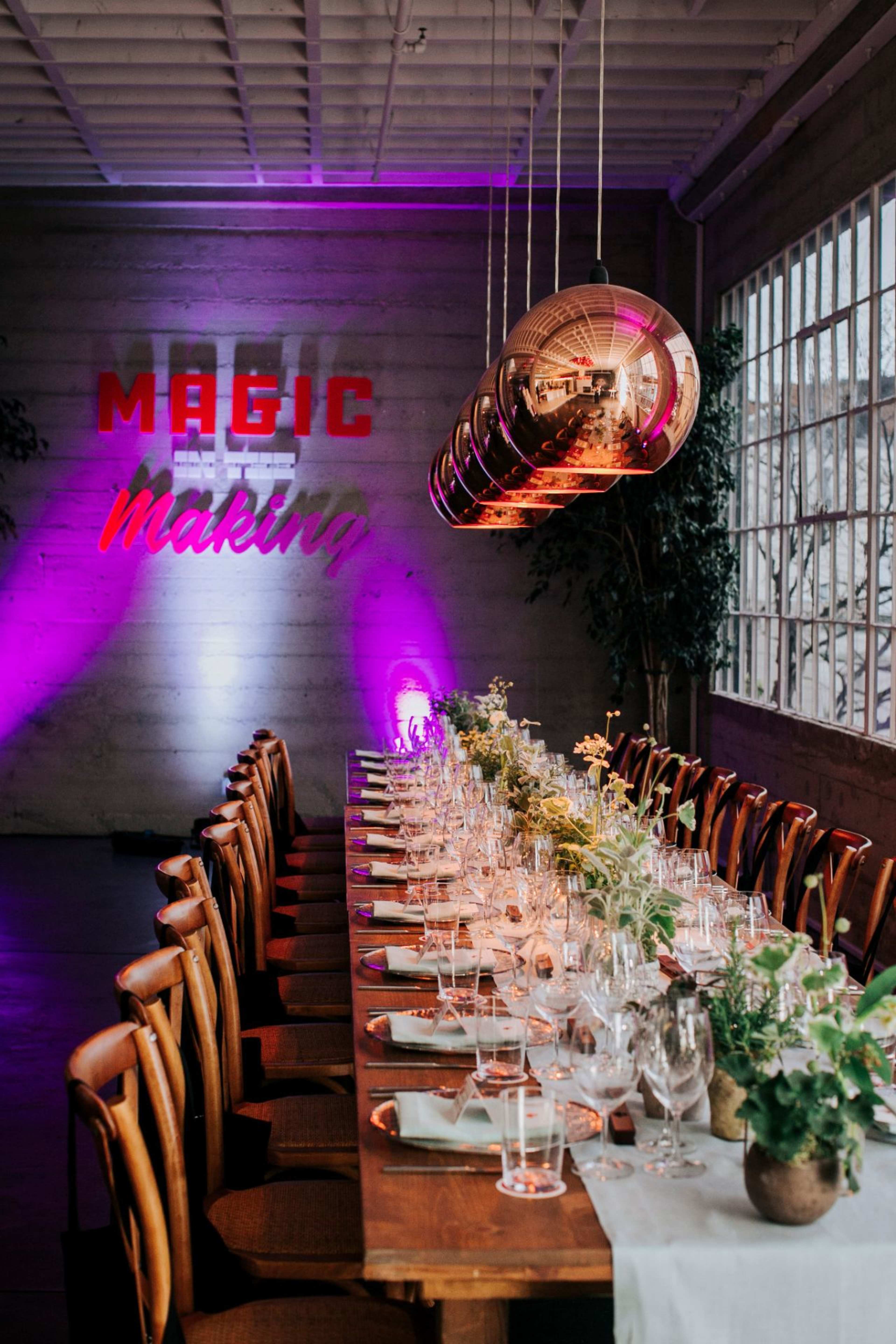 A long wooden dining table is elegantly set with glassware and greenery under modern pendant lights, with the words "MAGIC" and "Making" displayed on the wall in vibrant colors.
