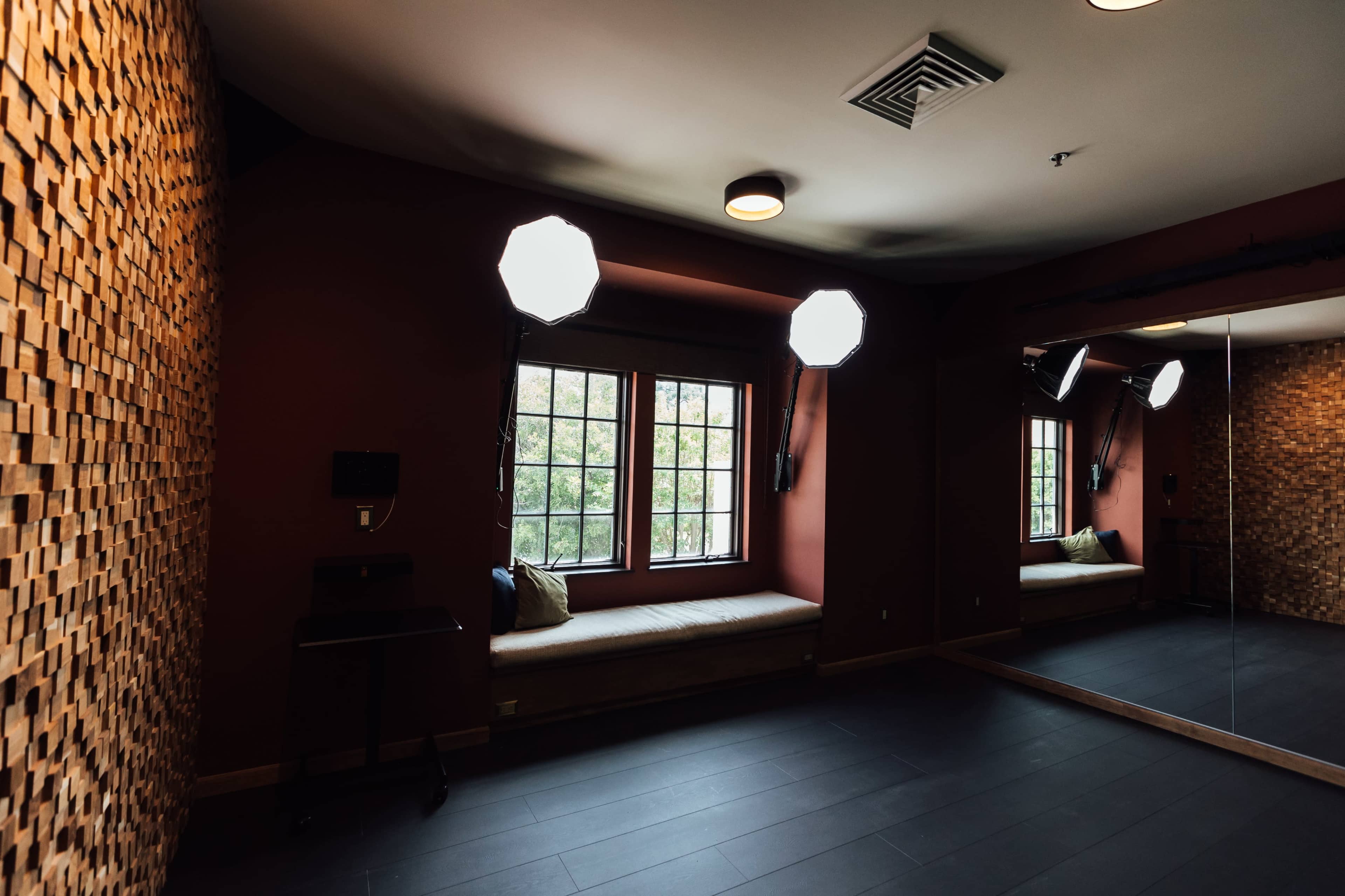 A soundproofed room with a wooden wall, large windows, and two softboxes for lighting, along with a mirror and a small table.