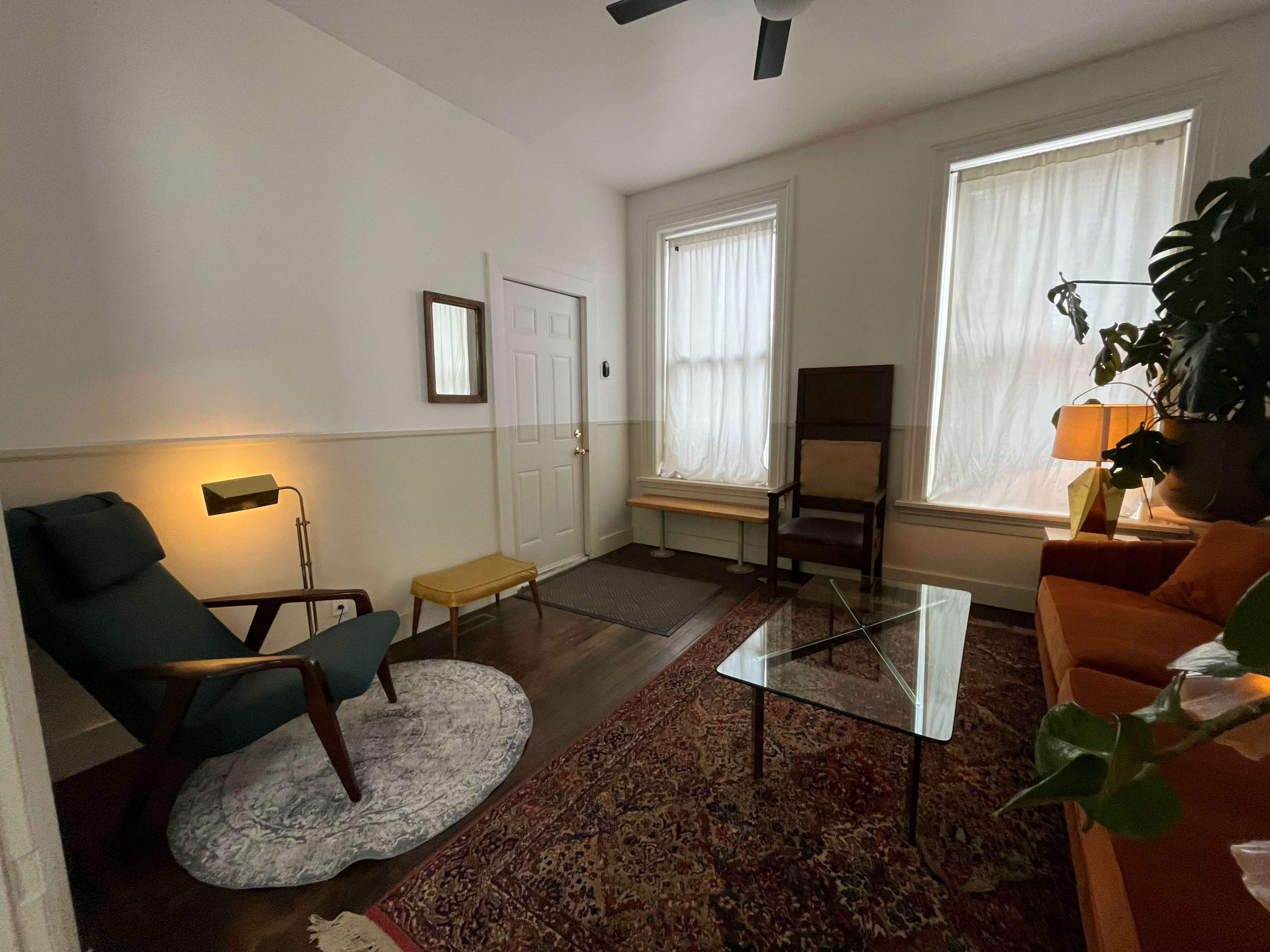 The room features a green armchair, a glass coffee table, an orange sofa, and a large window with sheer curtains.