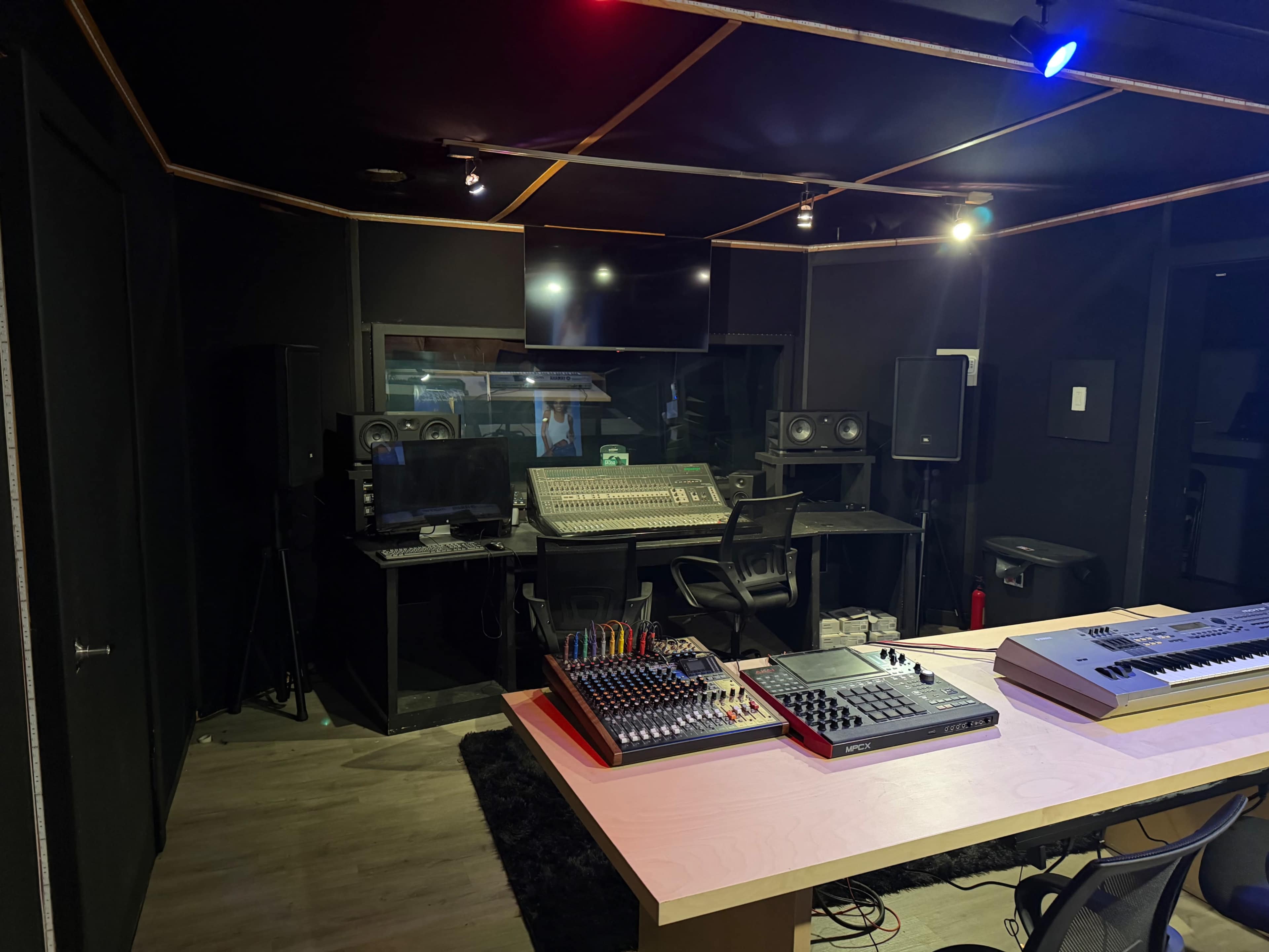 The image shows a modern music studio featuring a large mixing console, various electronic equipment on a table, and soundproofed walls.