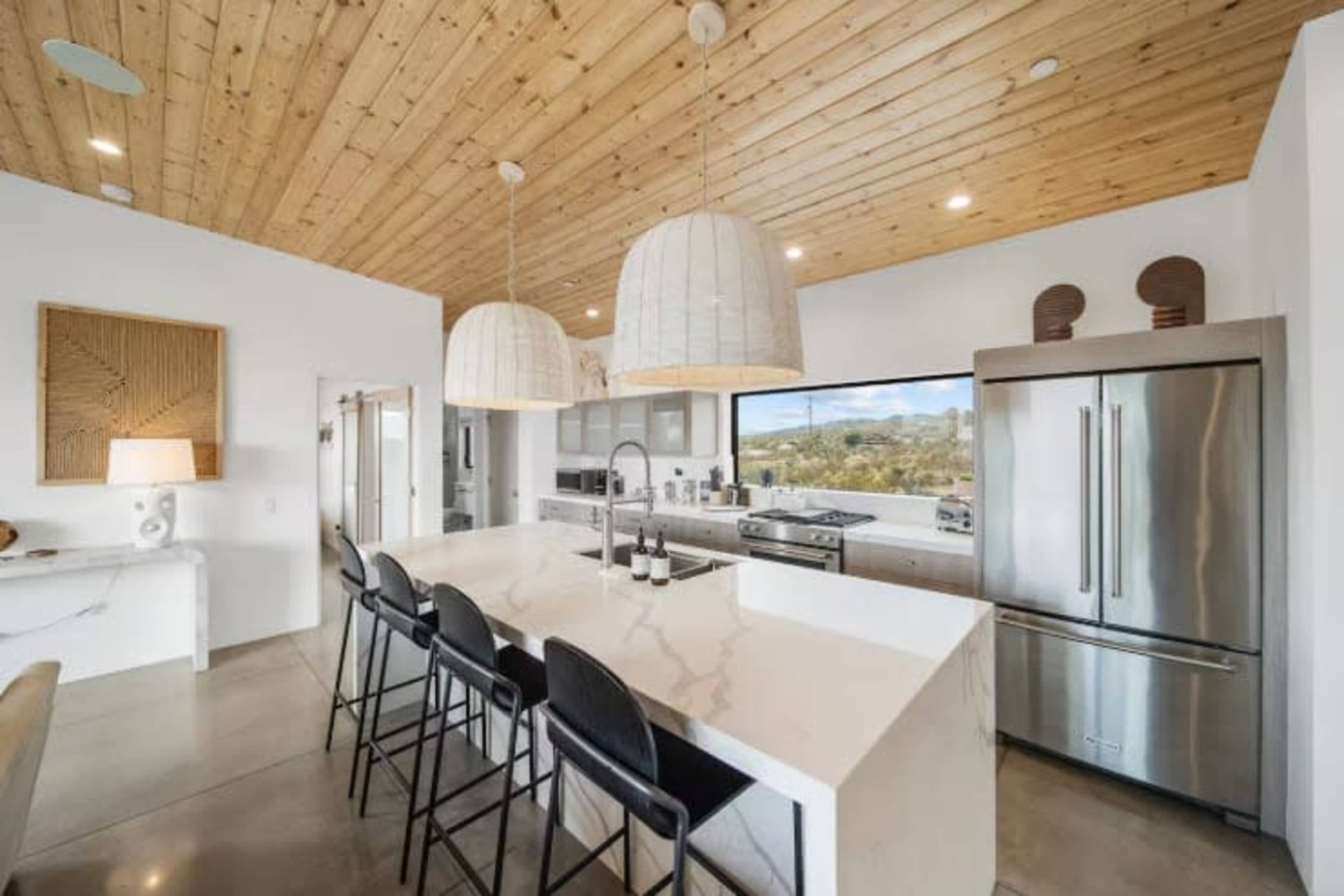 The kitchen features a large white marble island with black bar stools, pendant lighting, and a modern stainless steel refrigerator, all under a vaulted wooden ceiling.