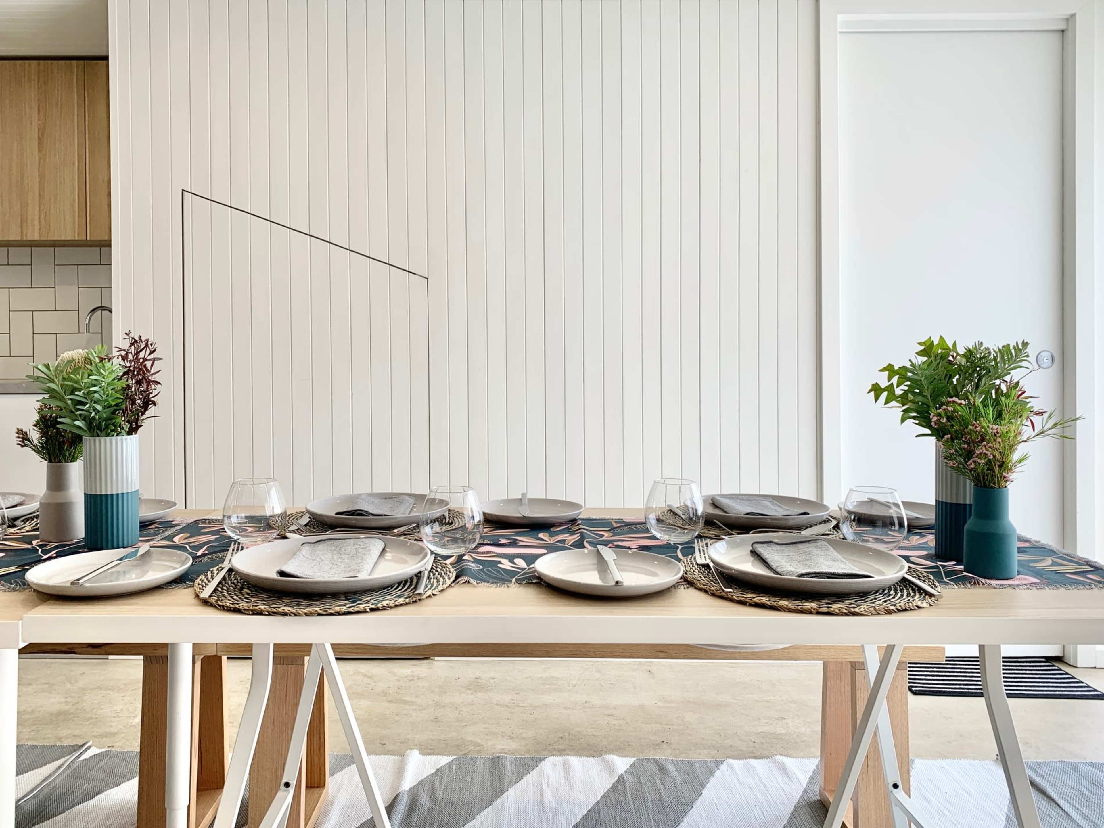 A dining table is set with plates, glasses, and decorative plants, against a backdrop of a modern kitchen and a white wall.