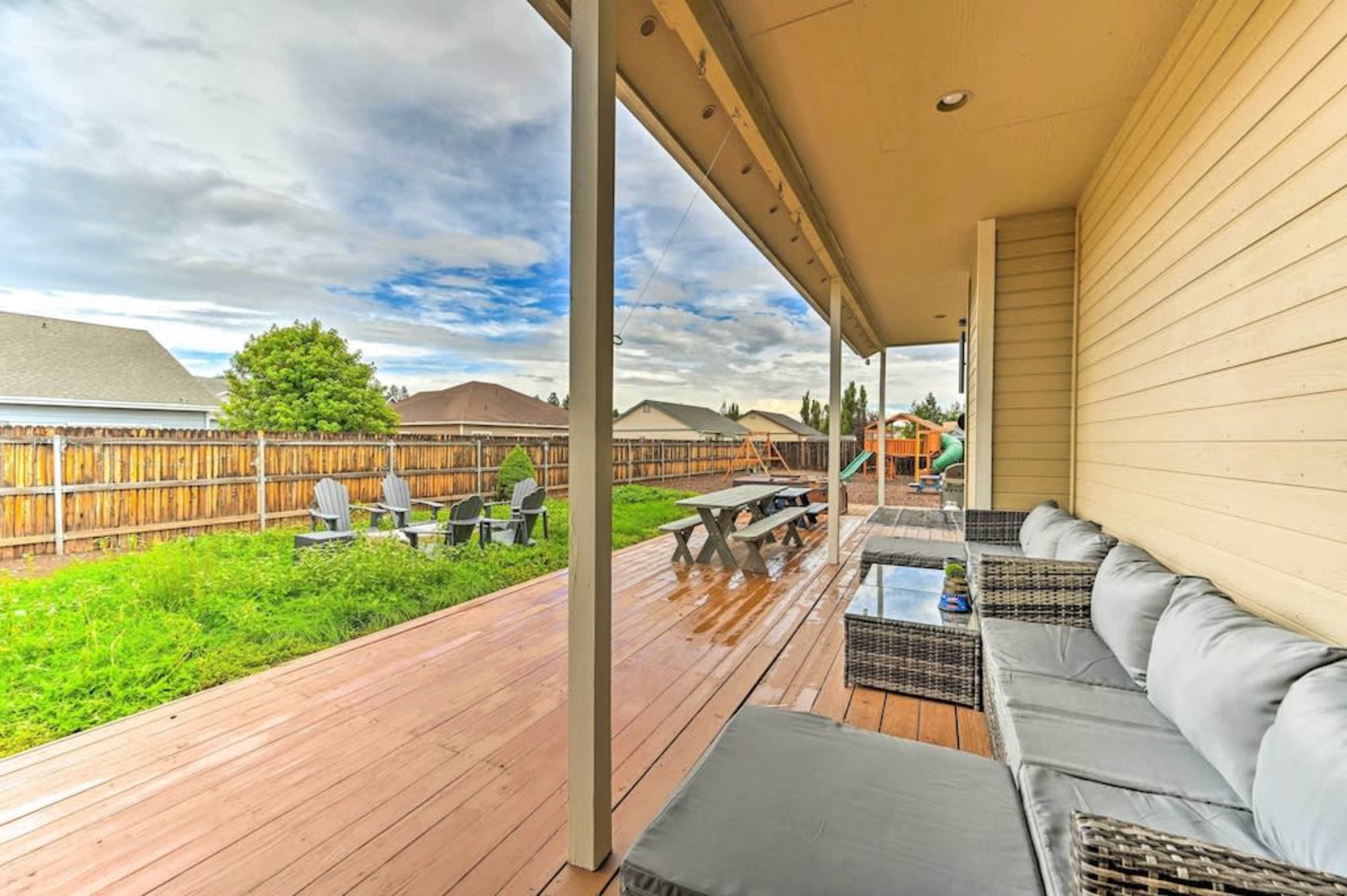A covered wooden deck features outdoor seating and dining areas, overlooking a grassy yard with a wooden fence in the background.