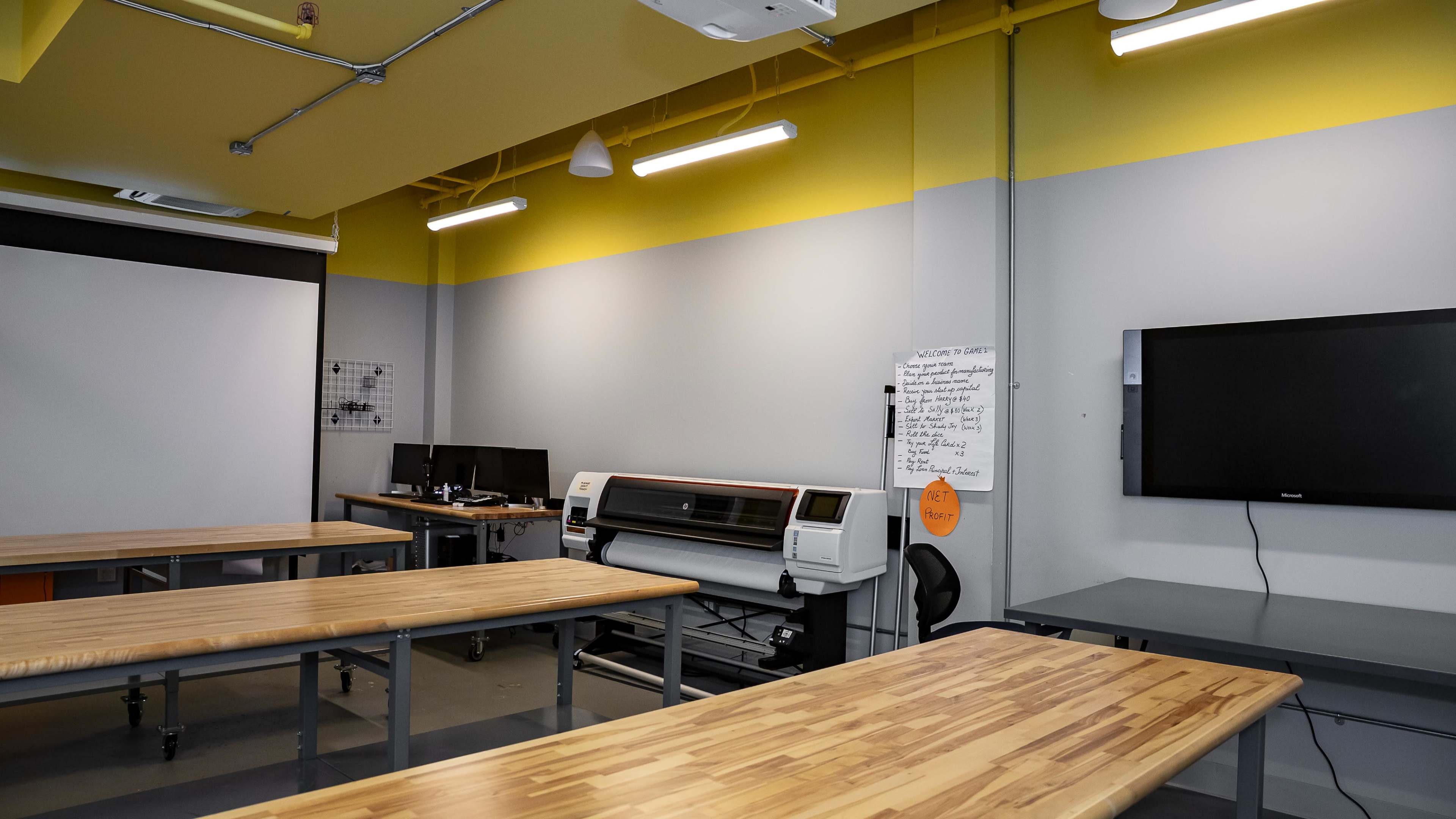 A spacious workshop area features wooden tables, a large printer, and electronic screens against a two-tone wall.