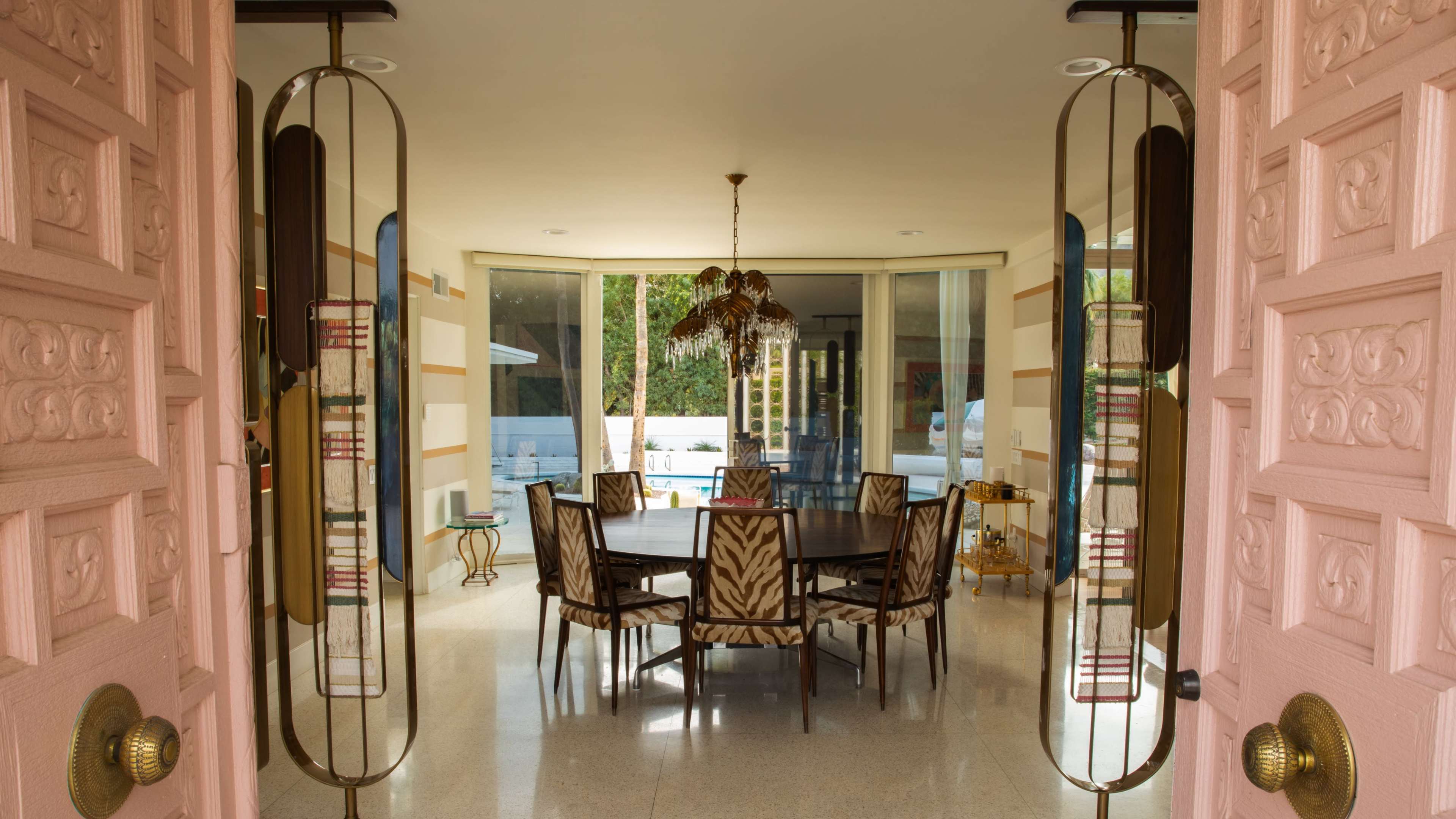 A large dining room with a round wooden table surrounded by patterned chairs is visible through double doors, leading to a space with large windows overlooking a pool.
