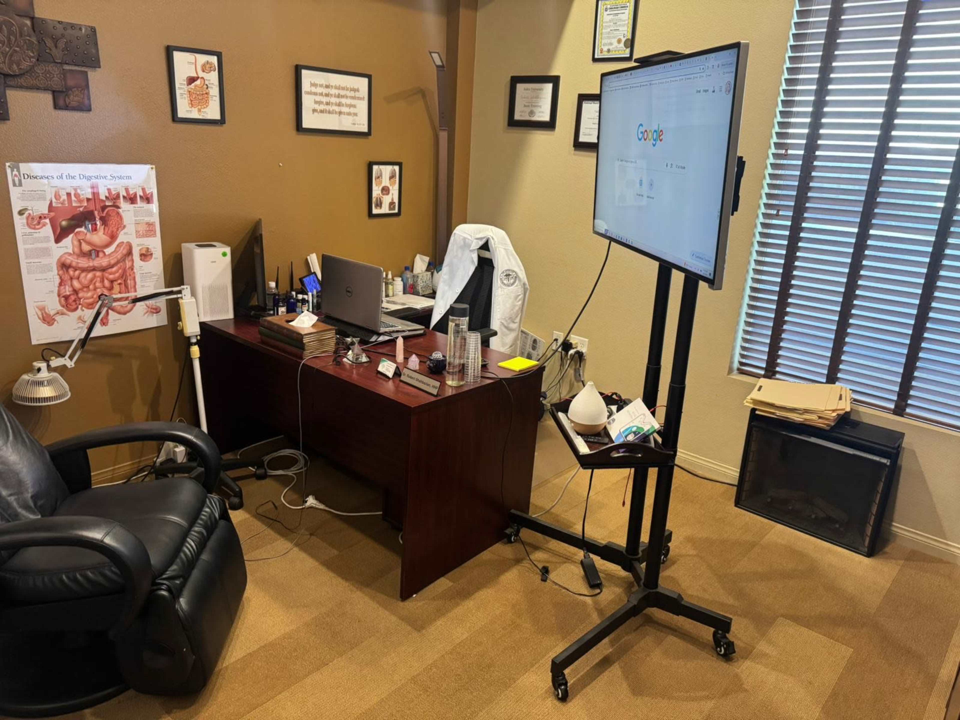 The image shows a medical office with a dark wooden desk, a computer monitor on a stand, medical textbooks, and a white coat hanging nearby.