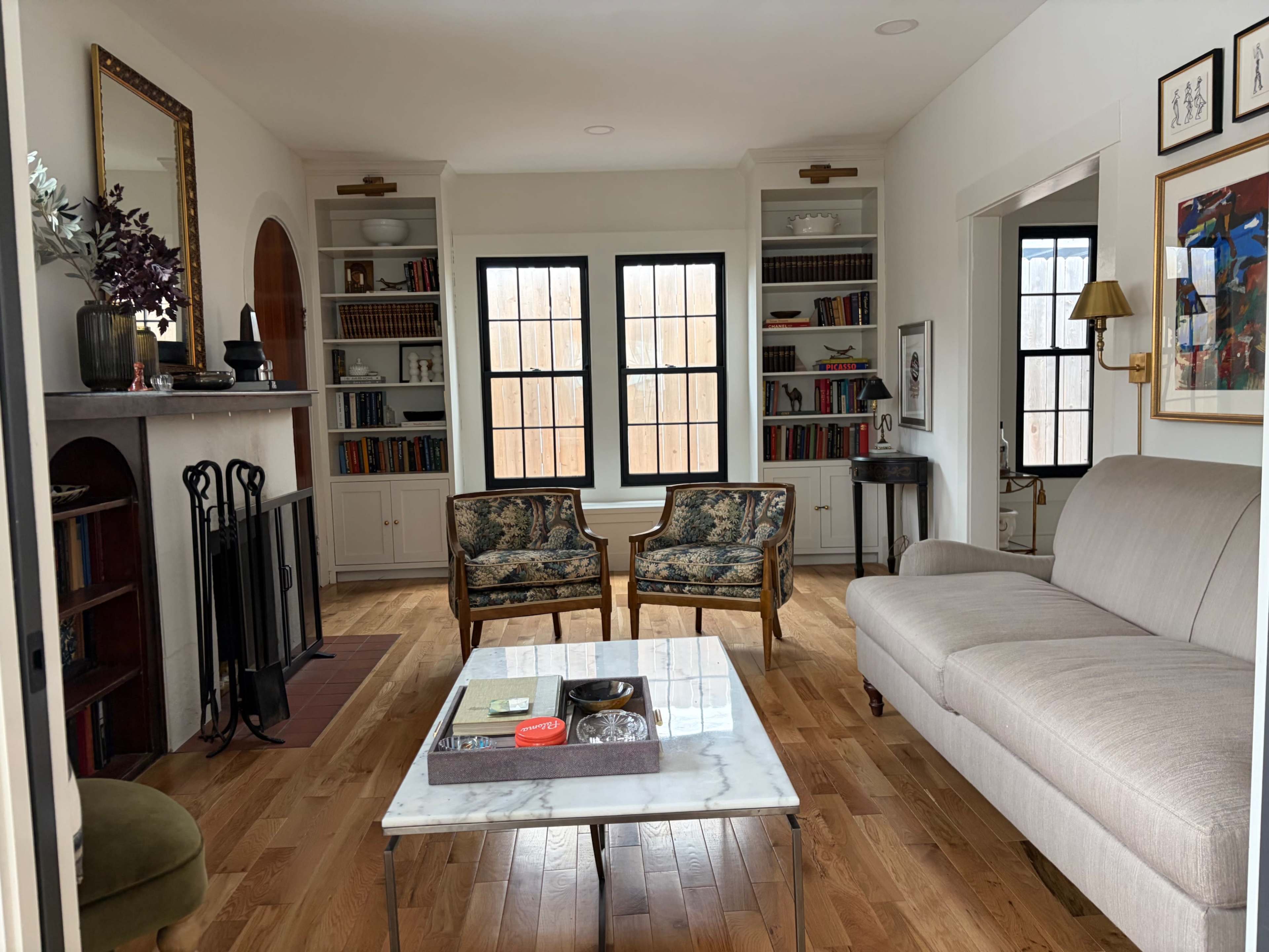 The image shows a living room with a fireplace, two patterned chairs, a sofa, a coffee table, and built-in bookshelves.