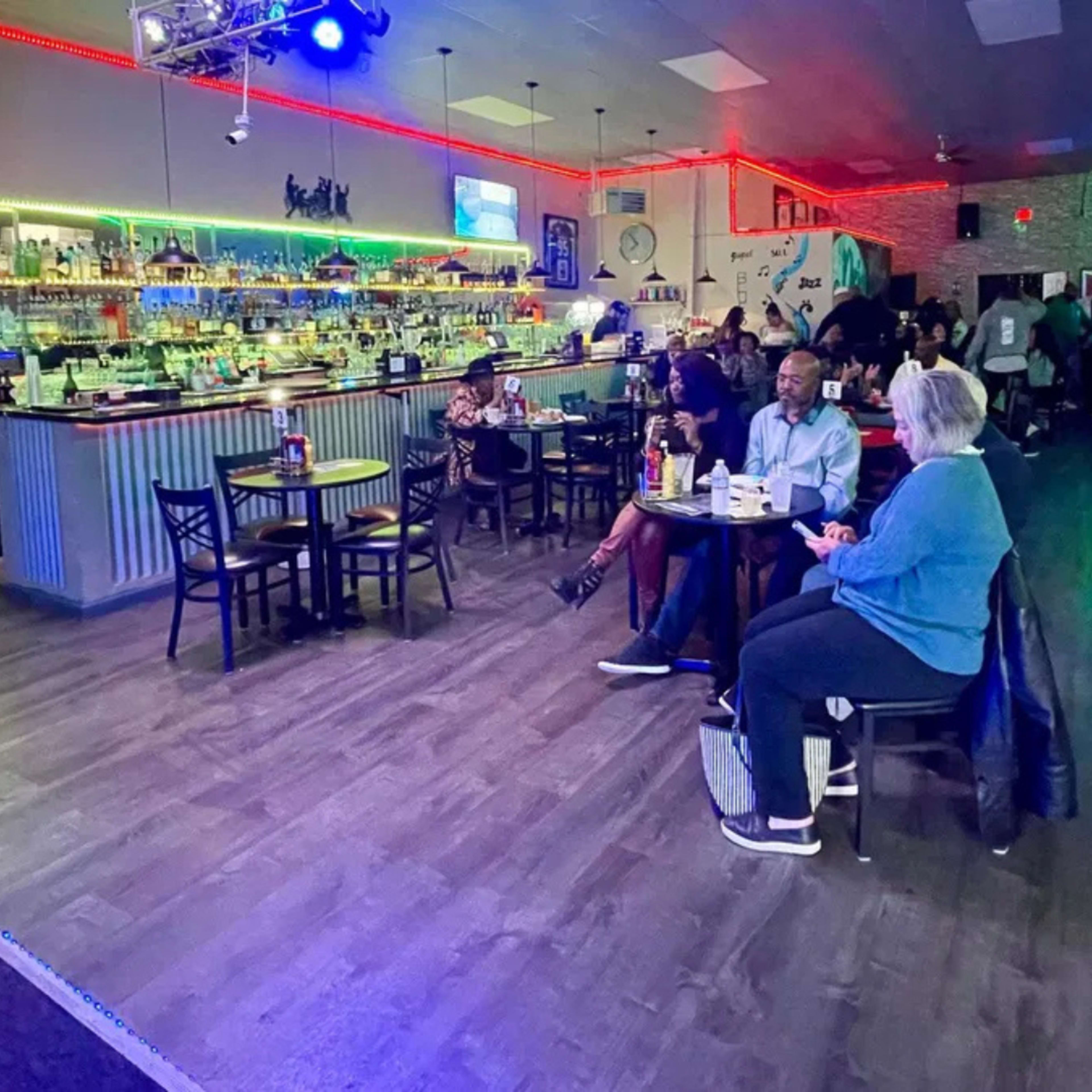 The image shows a dimly lit bar with a well-stocked backlit bar area and a few patrons seated at tables.