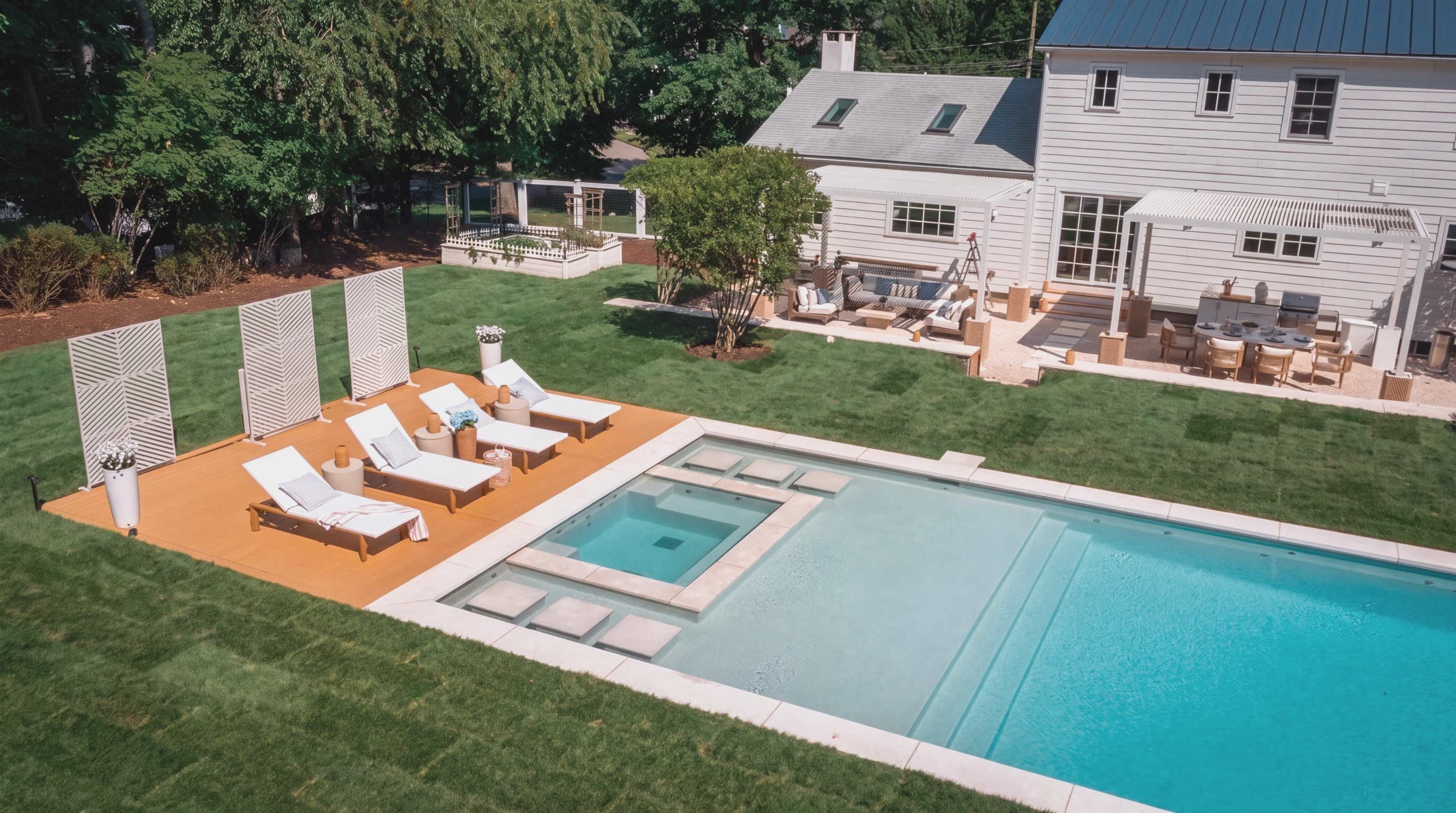 A modern backyard features a swimming pool with a spa area, surrounded by lounge chairs and landscaped greenery, alongside a white house with an outdoor dining space.
