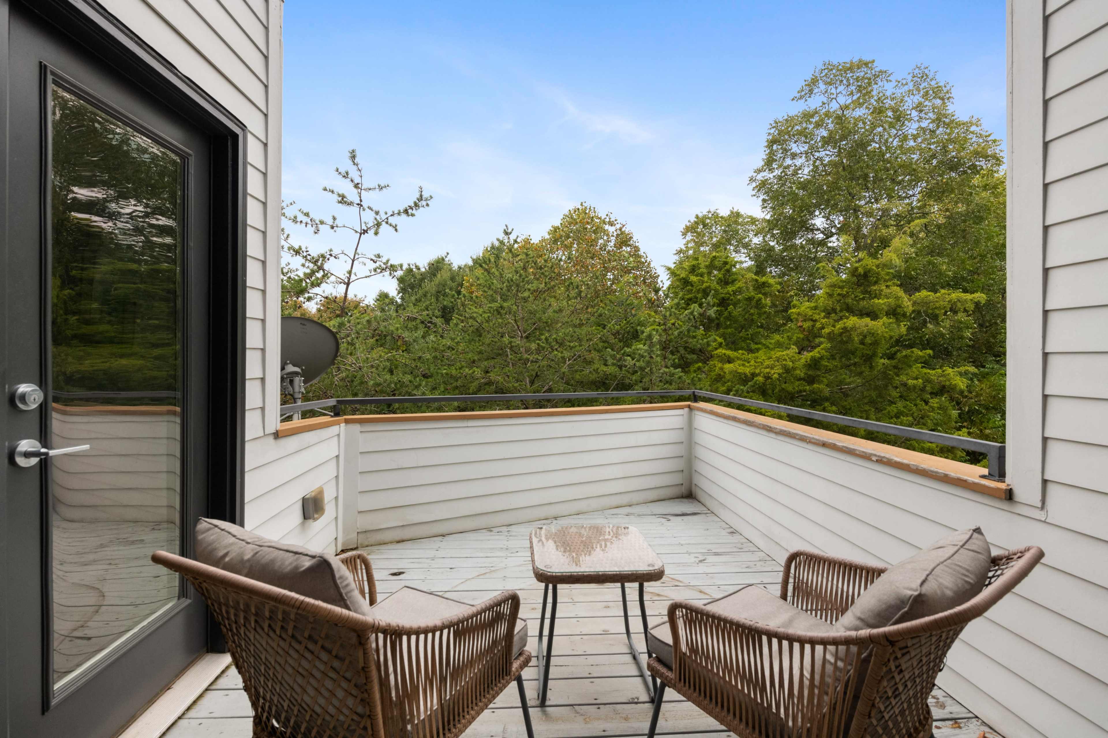 A small balcony features two wicker chairs and a table, surrounded by lush greenery and trees.