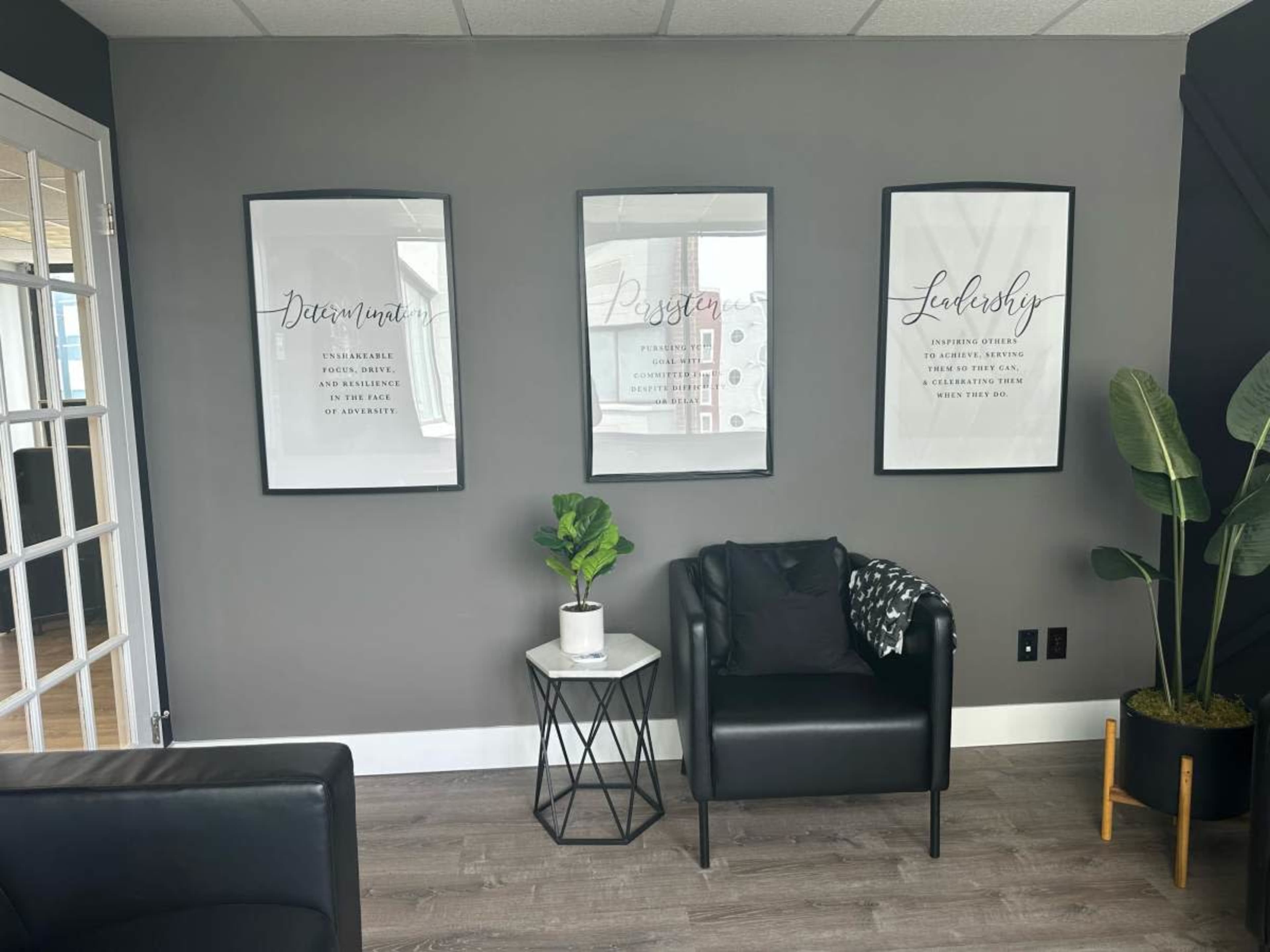 A modern office space with three framed motivational posters on the gray wall, a black armchair with a cushion, a small table, and a potted plant.
