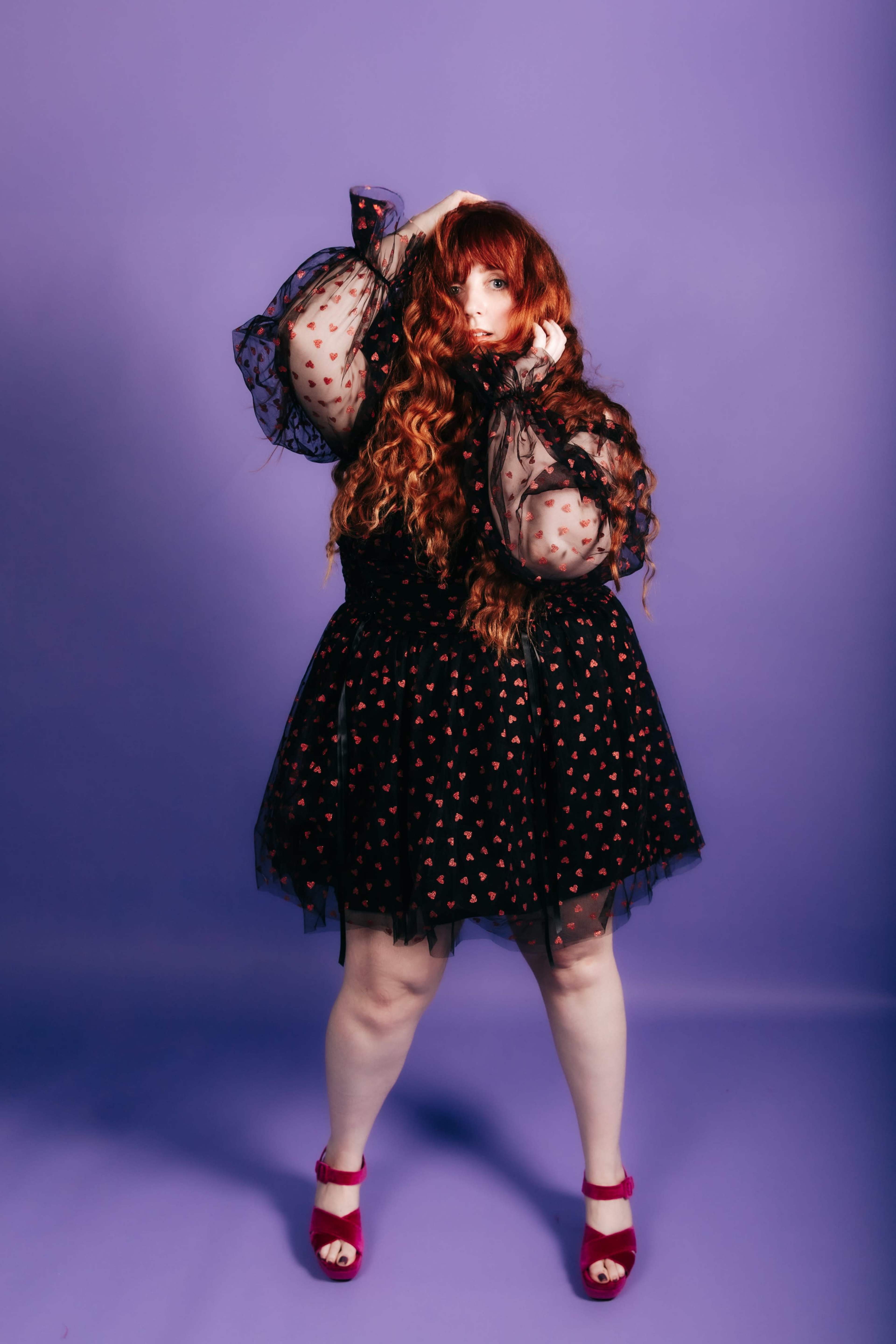 A person with long, wavy red hair poses in a black dress adorned with red hearts against a purple background.