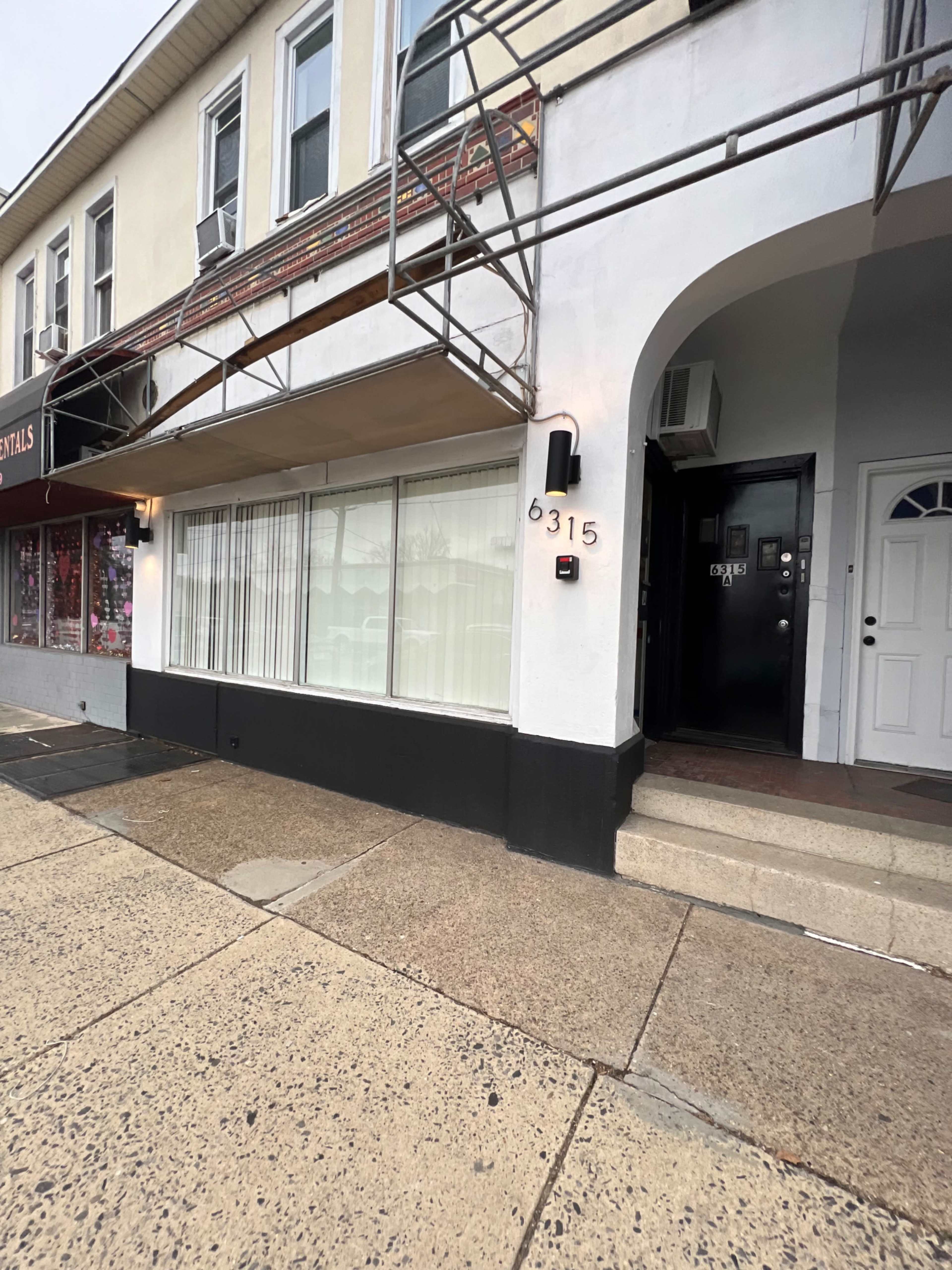 The image shows a building facade with a storefront featuring large windows, an entrance door on the right, and scaffolding above the sidewalk.