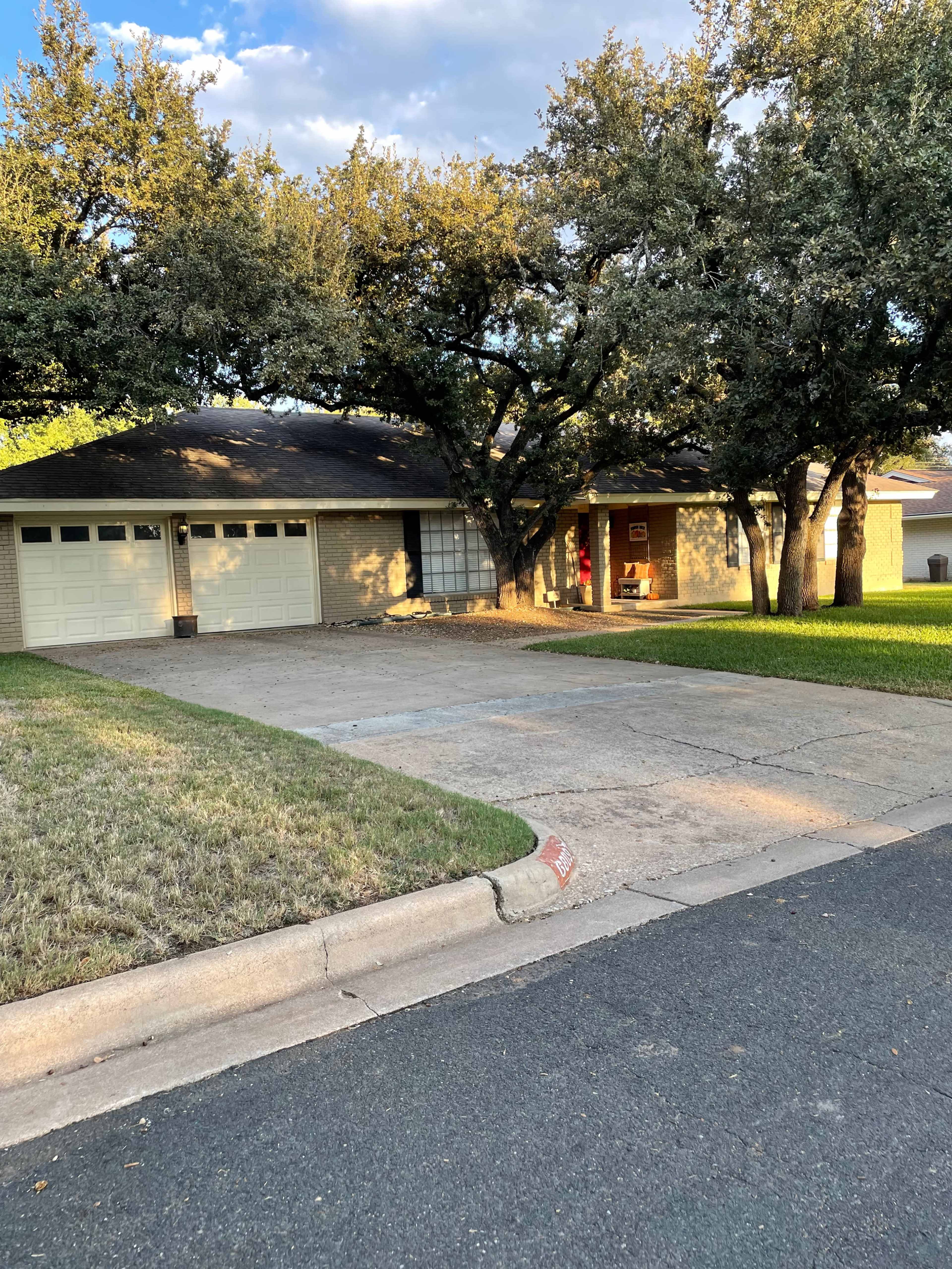 A single-story house with a two-car garage is set back from a quiet street, surrounded by large oak trees and a manicured lawn.