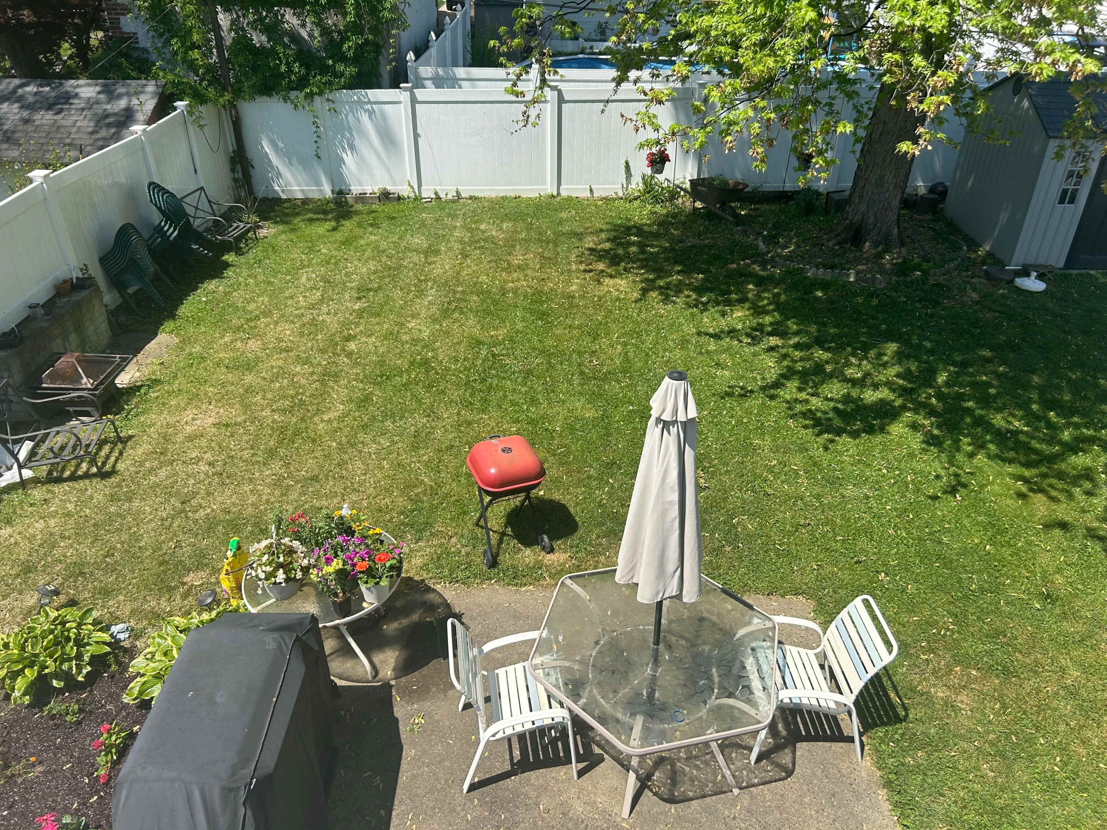 The image shows a backyard with a grassy area, a patio table with chairs, a grill, and flower pots, all enclosed by a white fence.